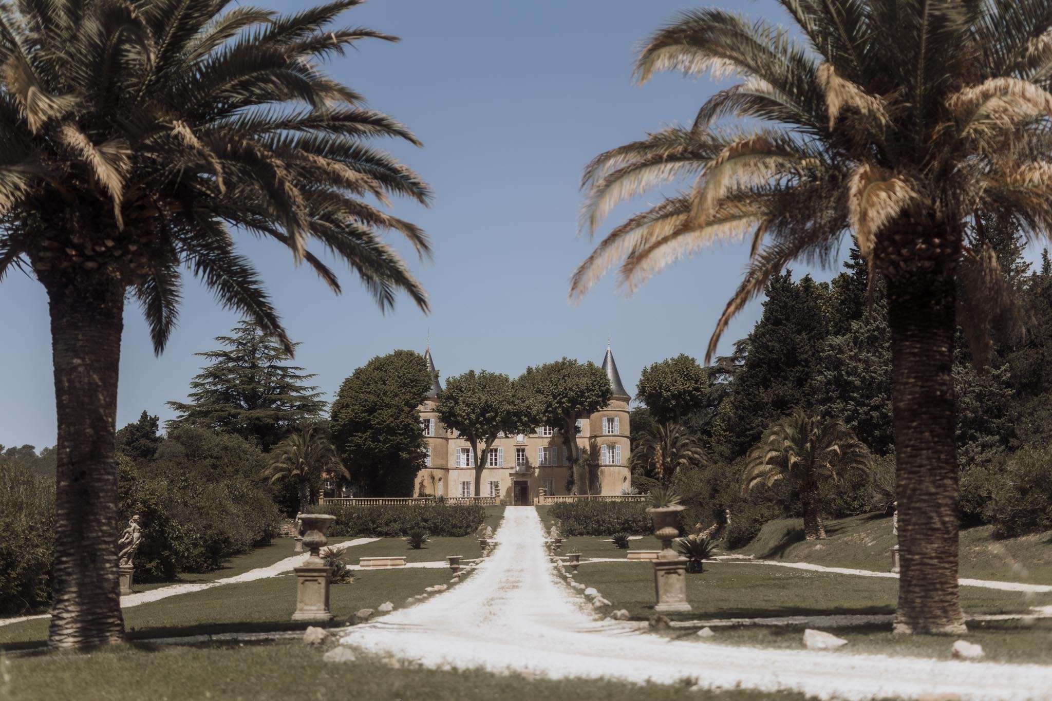 Symmetrical view down gravel driveway toward golden limestone chateau with blue-grey turrets and palm trees