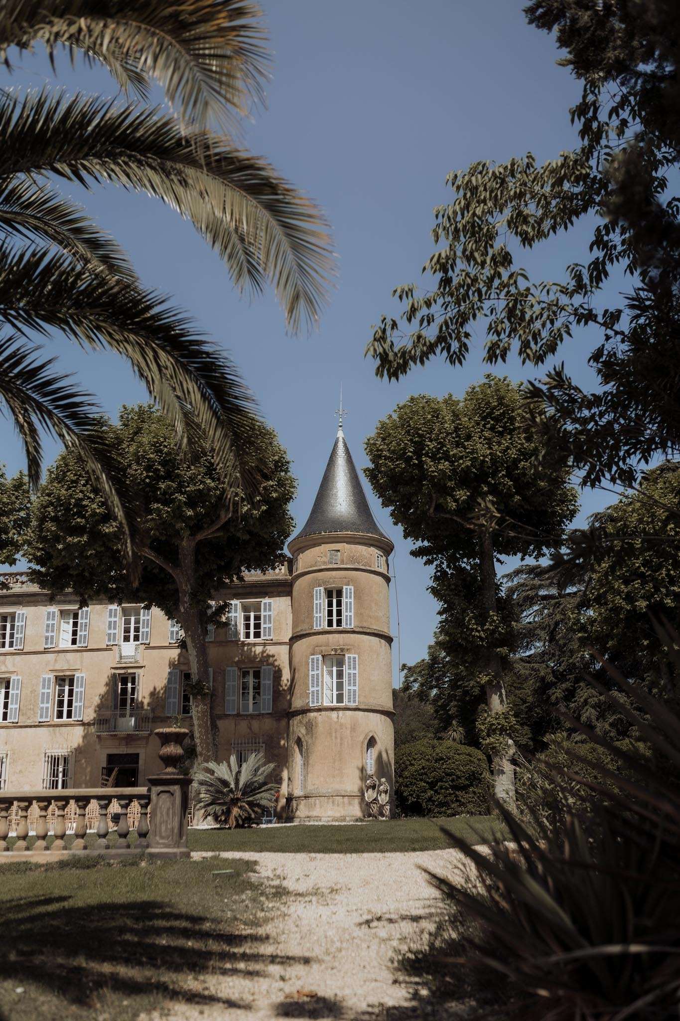 French chateau exterior with round tower, slate conical roof, golden stone facade, and white-shuttered windows framed by t...