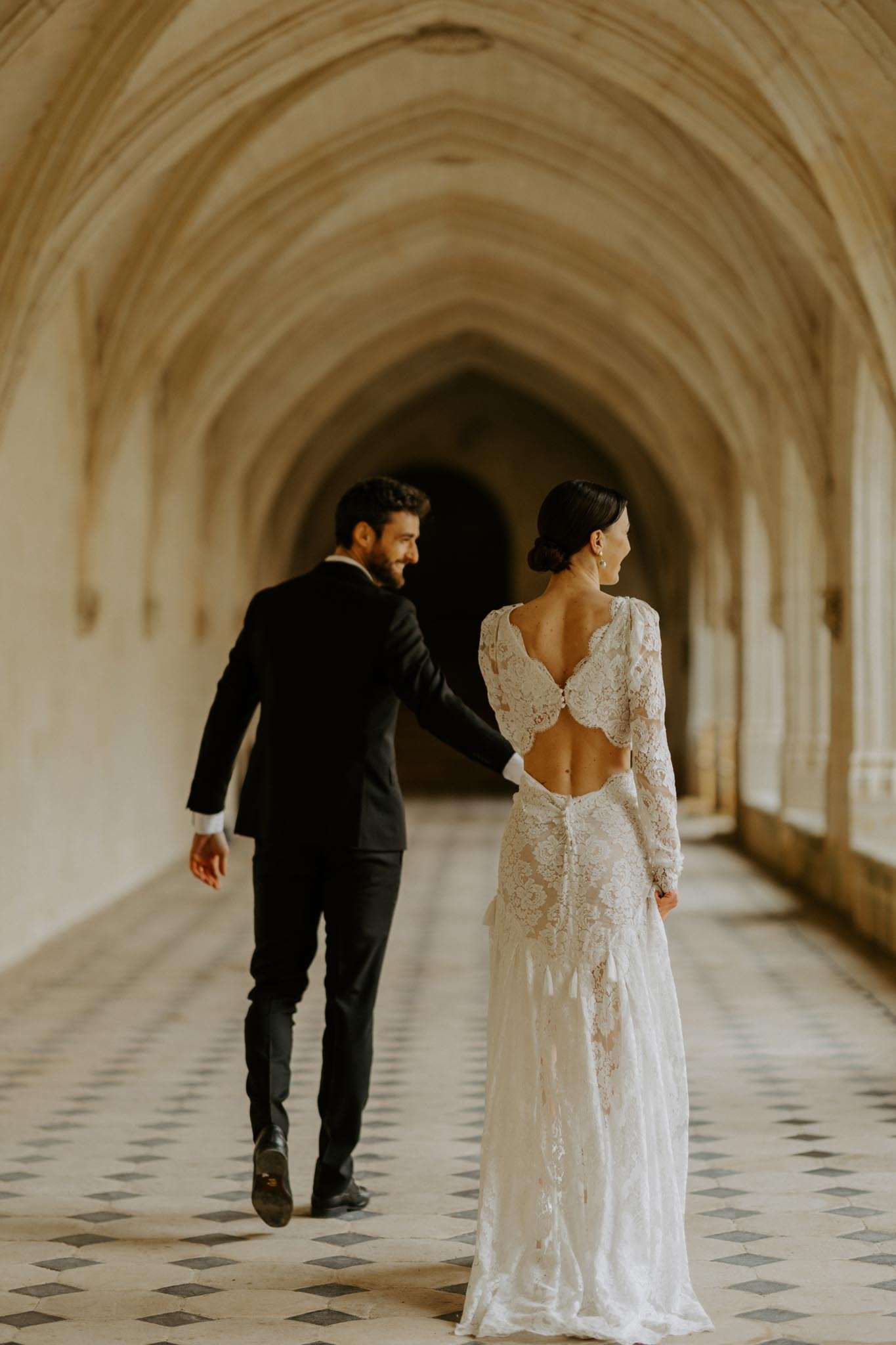 Couple walking through Gothic vaulted stone cloister bride in open-back lace gown groom in black suit diamond tile floor