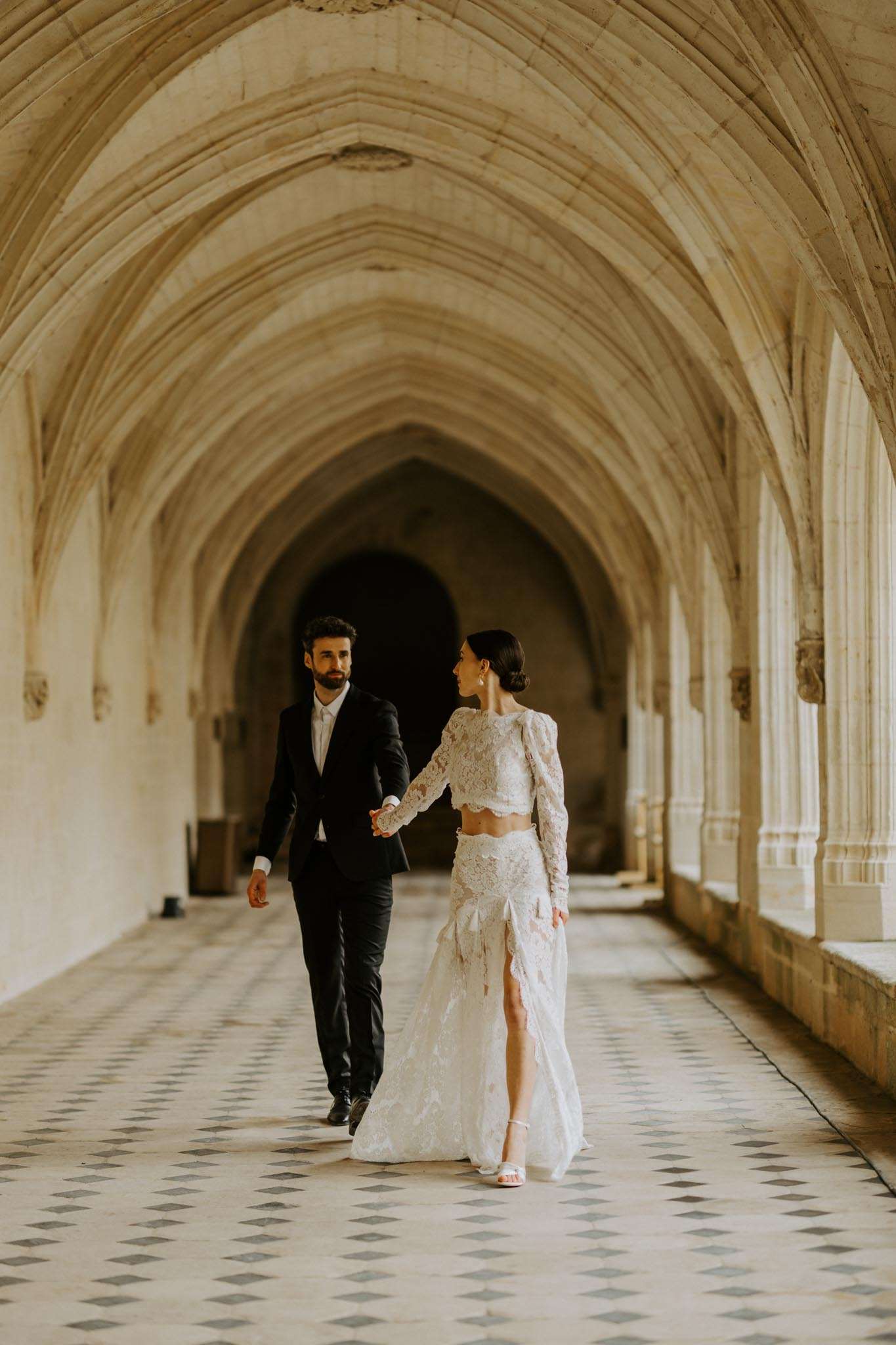 A couple portrait taken in the vaulted stone corridor of what appears to be a historic abbey or monastery, featuring Gothic ribbed ceiling arches and a diamond-patterned tile floor. The bride wears a two-piece ivory lace ensemble with long sleeves, a cropped top, and a high-slit skirt, paired with white heeled sandals and her dark hair in an updo; the groom wears a black suit with a white dress shirt and no tie. The two are walking hand-in-hand through the corridor, the bride turning back to look at the groom, captured in a full-length portrait composition with the arched hallway receding into the background. The styling is modern with a classic architectural backdrop. Potential venue feature image.