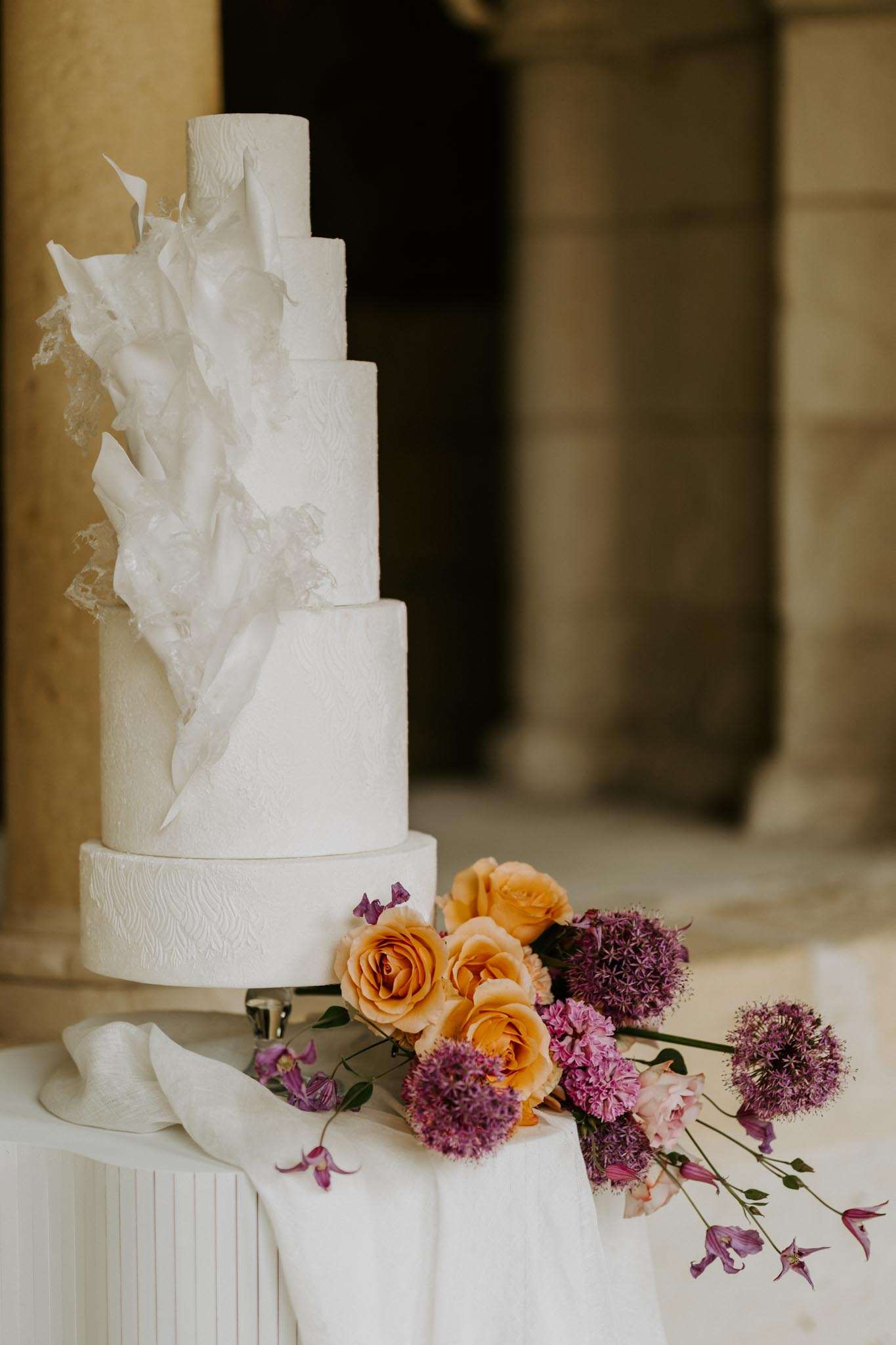 Close-up detail shot of a tall five-tier white wedding cake displayed on a white linen-covered pedestal table, set against the stone columns of what appears to be a classical French château or manor exterior. The cake features an all-white textured finish with an architectural sugar work design — large, irregular white sugar or wafer paper shards cascading down one side alongside what appears to be lace-like sugar detailing. At the base of the cake, a loose arrangement of amber/apricot garden roses, purple allium globes, hot pink carnations, blush roses, and trailing purple clematis blooms is casually placed on the table alongside a draped white silk ribbon. The overall styling is modern and sculptural with a bold floral color palette contrasting against the all-white cake.
