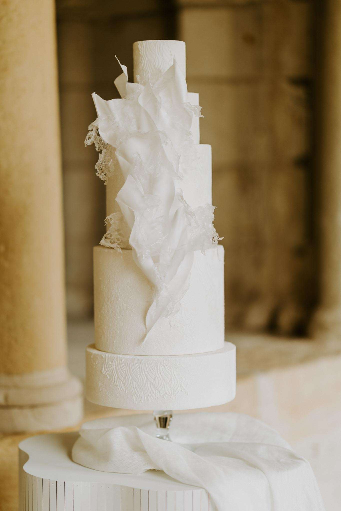 Five-tier all-white cake with draped wafer-paper sculpture on crystal pedestal before stone columns