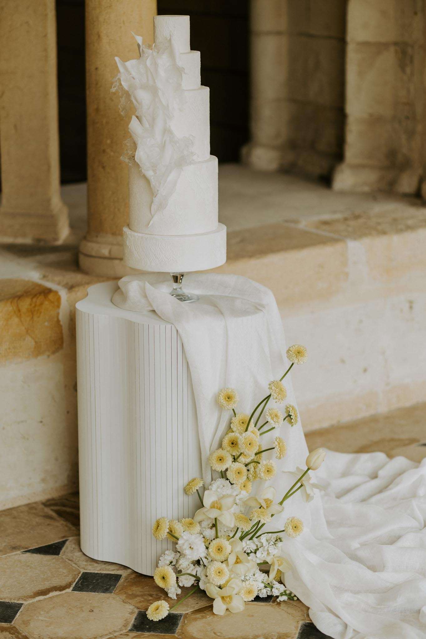 Five-tier white wedding cake with sculptural ruffles on fluted pedestal with yellow and white flowers in stone colonnade
