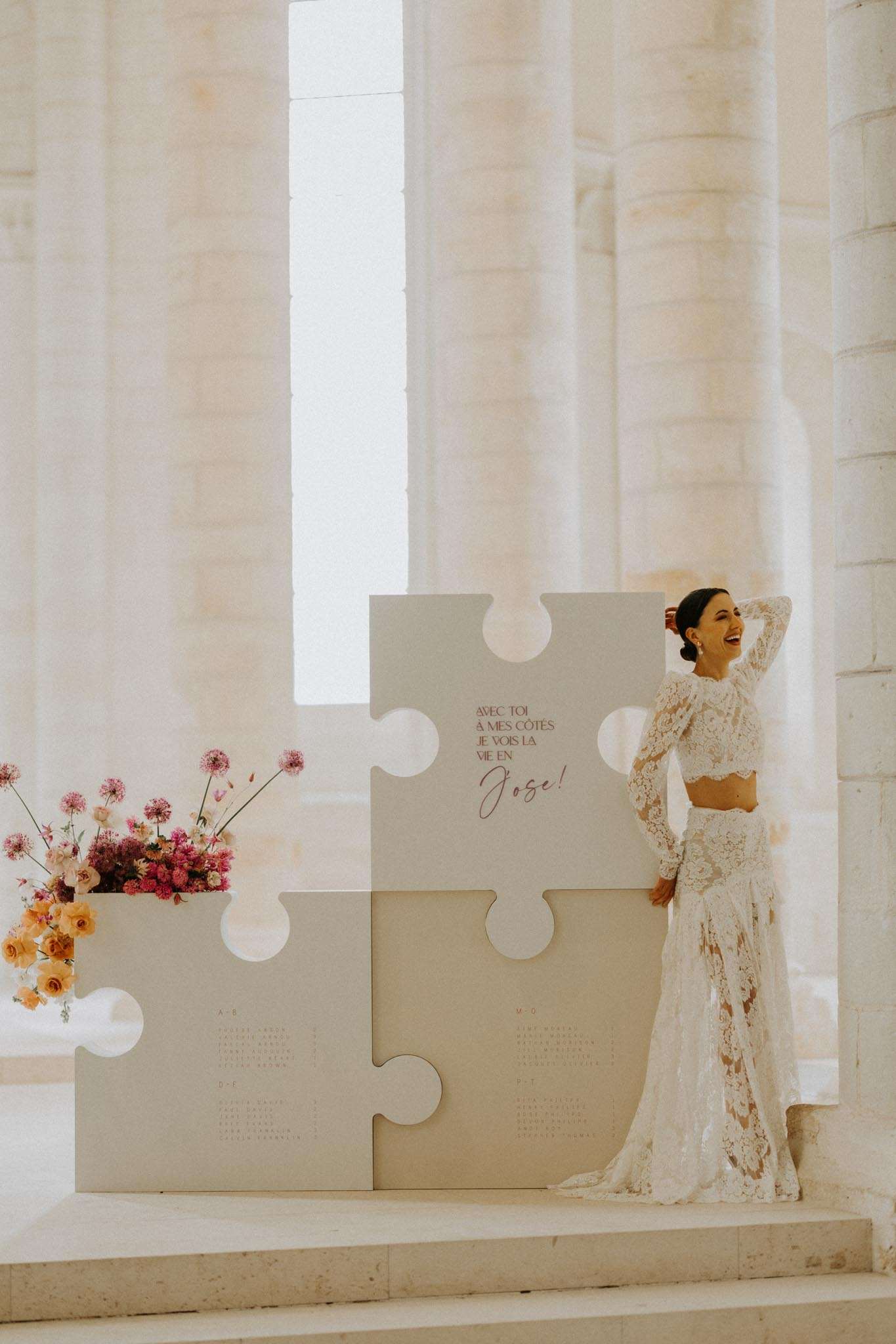 A bride stands laughing beside an oversized decorative puzzle-piece seating chart installation inside what appears to be a historic stone chapel or abbey with tall pale limestone columns and arched windows. The seating chart panels are cream-colored and shaped as interlocking puzzle pieces, with French text reading 'Avec toi à mes côtés je vois la vie en rose!' printed in pink script on the central piece. The bride wears a two-piece ivory lace gown consisting of a long-sleeve crop top and a fitted fishtail skirt with allover floral lace detailing. To the left of the installation sits a floral arrangement featuring hot pink scabiosa, magenta ranunculus, orange ranunculus, and long-stemmed dried or wildflowers in warm tones. The overall decor palette combines cream, blush, and warm orange-pink tones in a modern, editorial style. Full-length portrait shot.