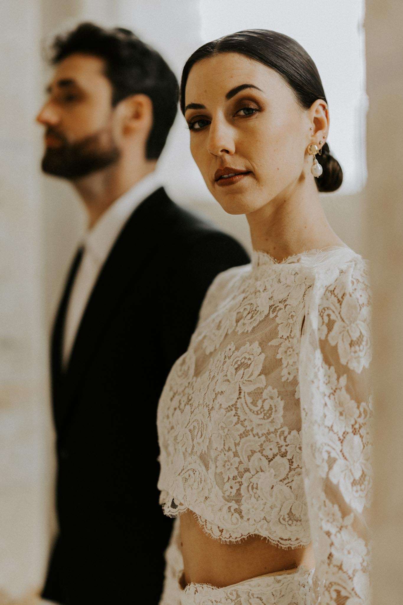 A close-up portrait of a bride in the foreground with the groom softly out of focus behind her, shot indoors against a light neutral background. The bride wears a two-piece ivory lace bridal outfit featuring a long-sleeved crop top with floral lace detailing and a high neckline, paired with a matching lace skirt; her dark hair is pulled back into a low sleek bun and she wears gold and pearl drop earrings. The groom is dressed in a black suit with a white shirt. The composition uses a shallow depth of field to draw focus to the bride's face and lace top details, giving the image a modern, pared-back aesthetic.