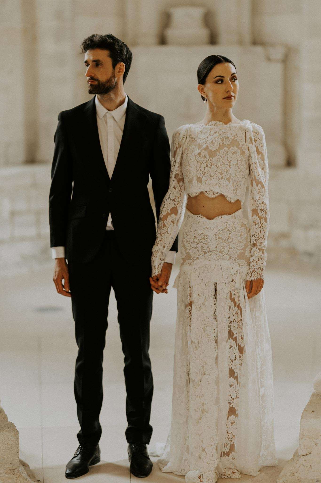 Bride in two-piece white lace bridal set and groom in black suit standing side by side against carved stone columns