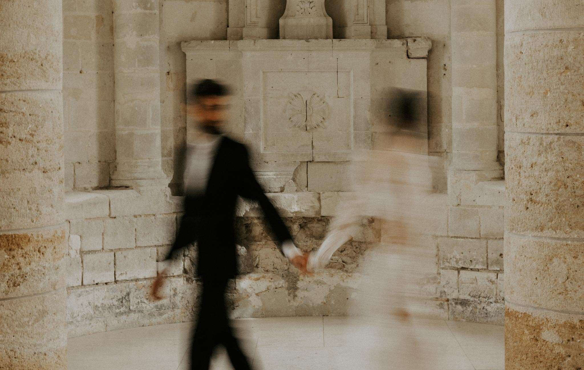 Motion-blurred couple walking through limestone arcade with carved stone panels at French abbey