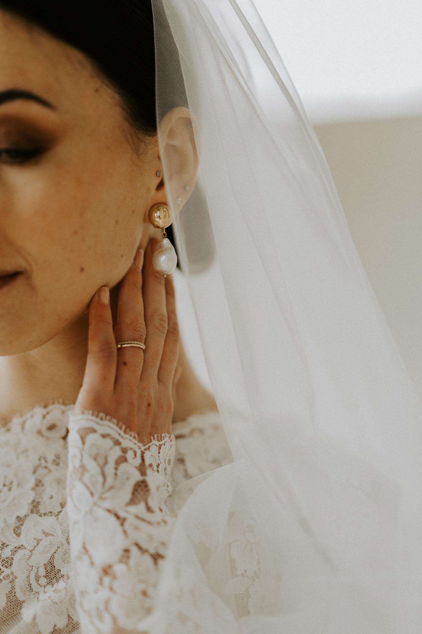 Close-up of bride with eyes closed wearing ivory lace sleeve, tulle veil, and gold baroque pearl drop earring