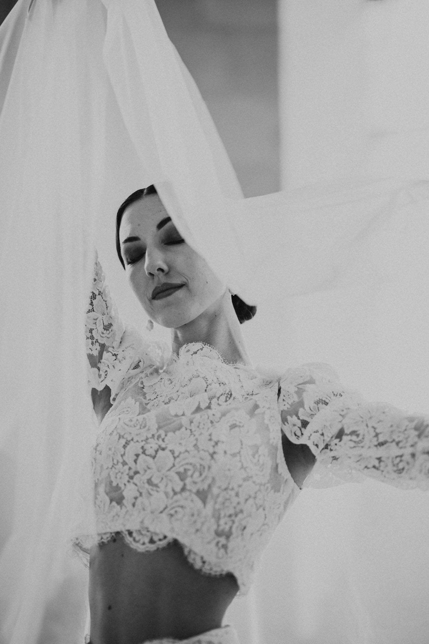 Black-and-white low-angle portrait of bride with cathedral veil billowing above her and cropped lace bridal top