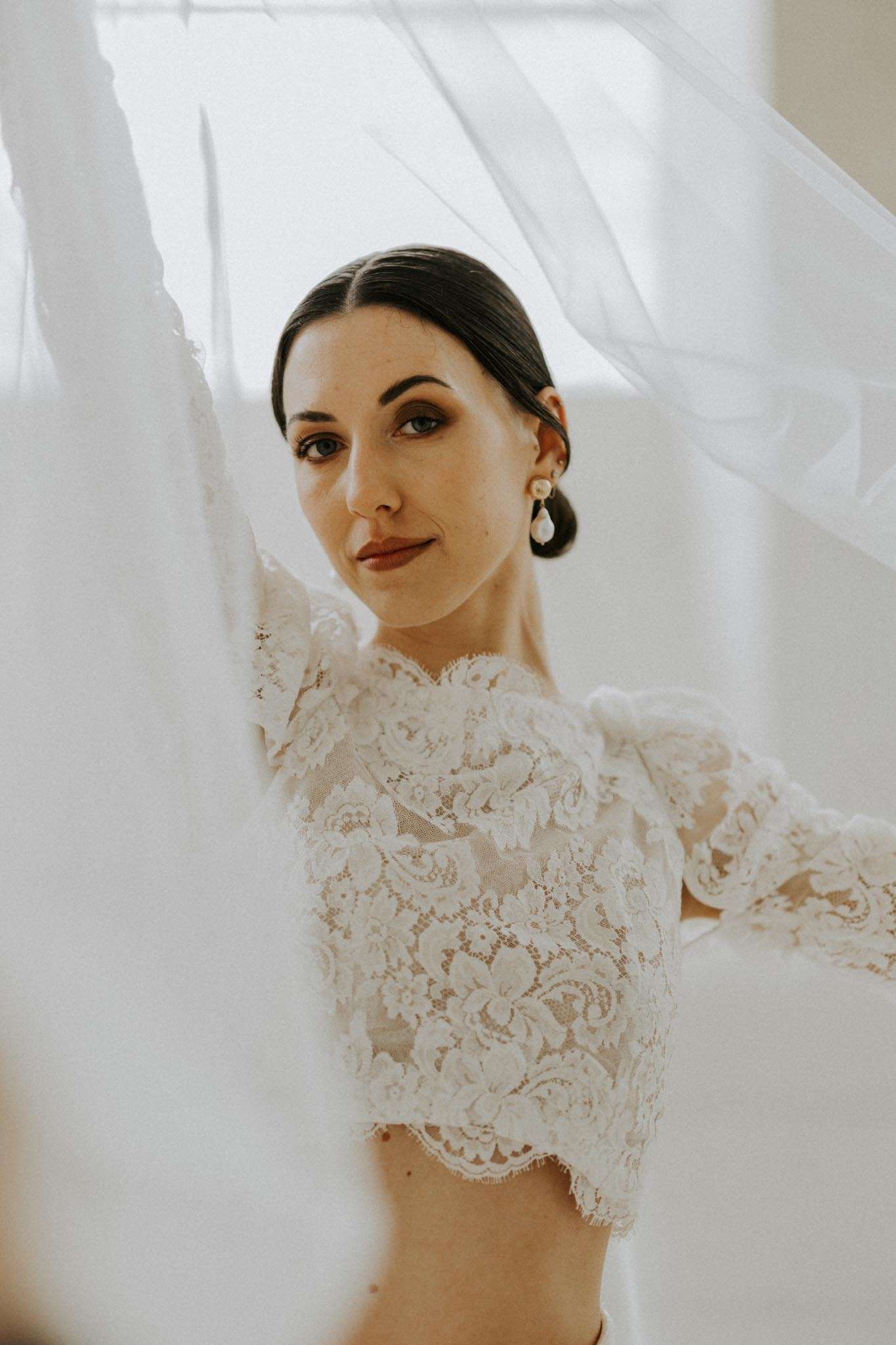 Bride holding tulle veil framing her face wearing ivory lace crop top with pearl earrings and sleek bun
