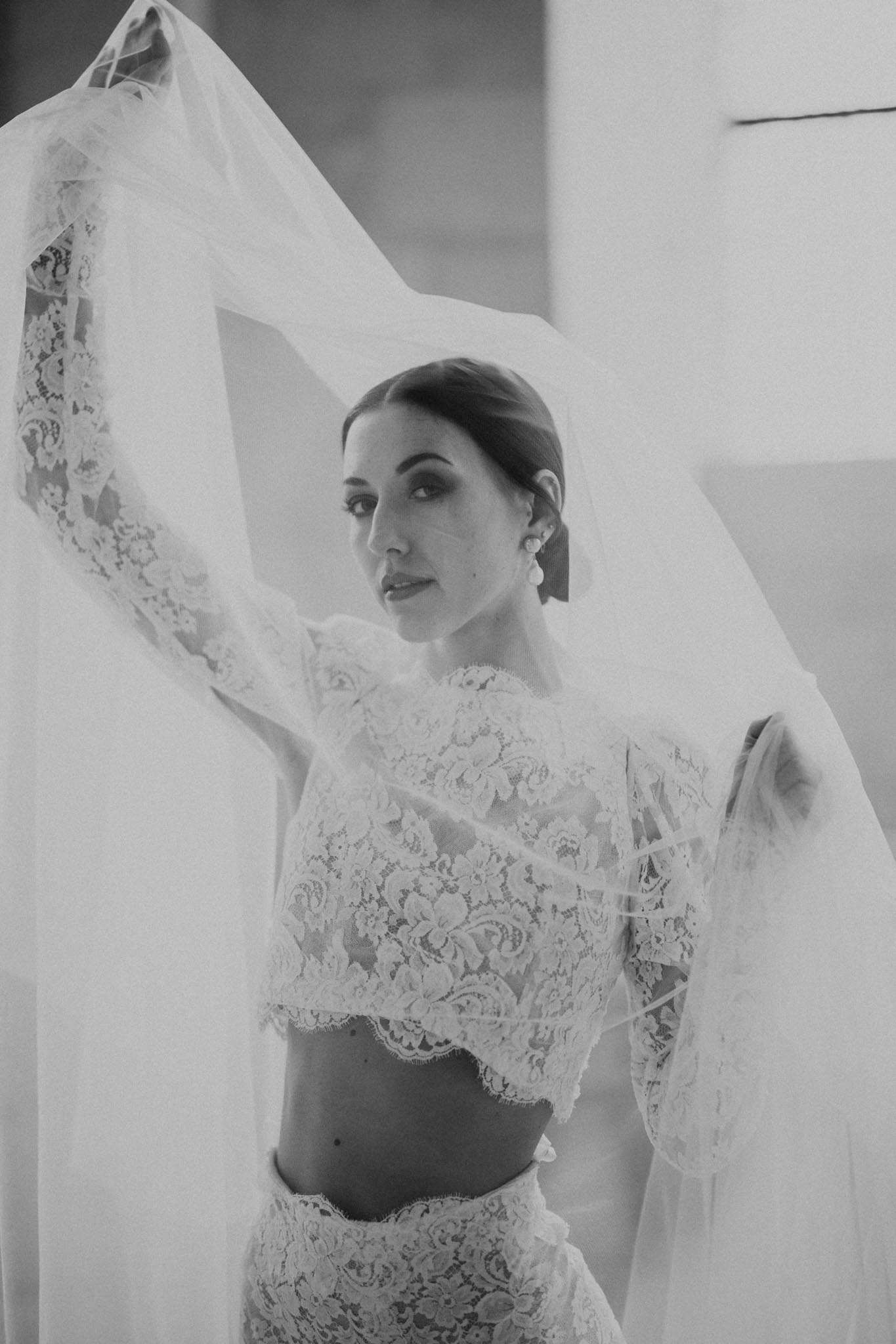 This is a black-and-white bridal portrait of a bride standing indoors against a plain light-coloured wall. She is wearing a two-piece lace bridal outfit consisting of a cropped long-sleeve lace top with a scalloped hem and a matching high-waisted lace skirt, with her midriff visible. She holds her long tulle veil above her head with both arms raised, allowing it to billow and drape around her. Her dark hair is swept back in a low updo, and she wears statement pearl or crystal drop earrings with defined eye makeup. The image is shot in high contrast black-and-white with soft, bright highlights on the veil and mid-tones on her skin. The composition is a close three-quarter portrait with a slightly low angle, giving a dynamic, editorial feel.