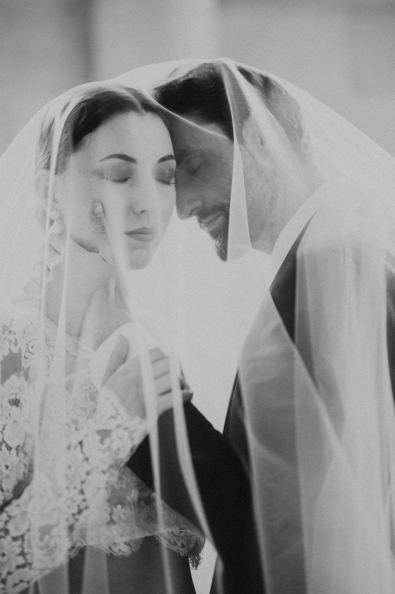 Bride and groom touch foreheads under draped veil with lace sleeve detail visible in soft B&W close-up