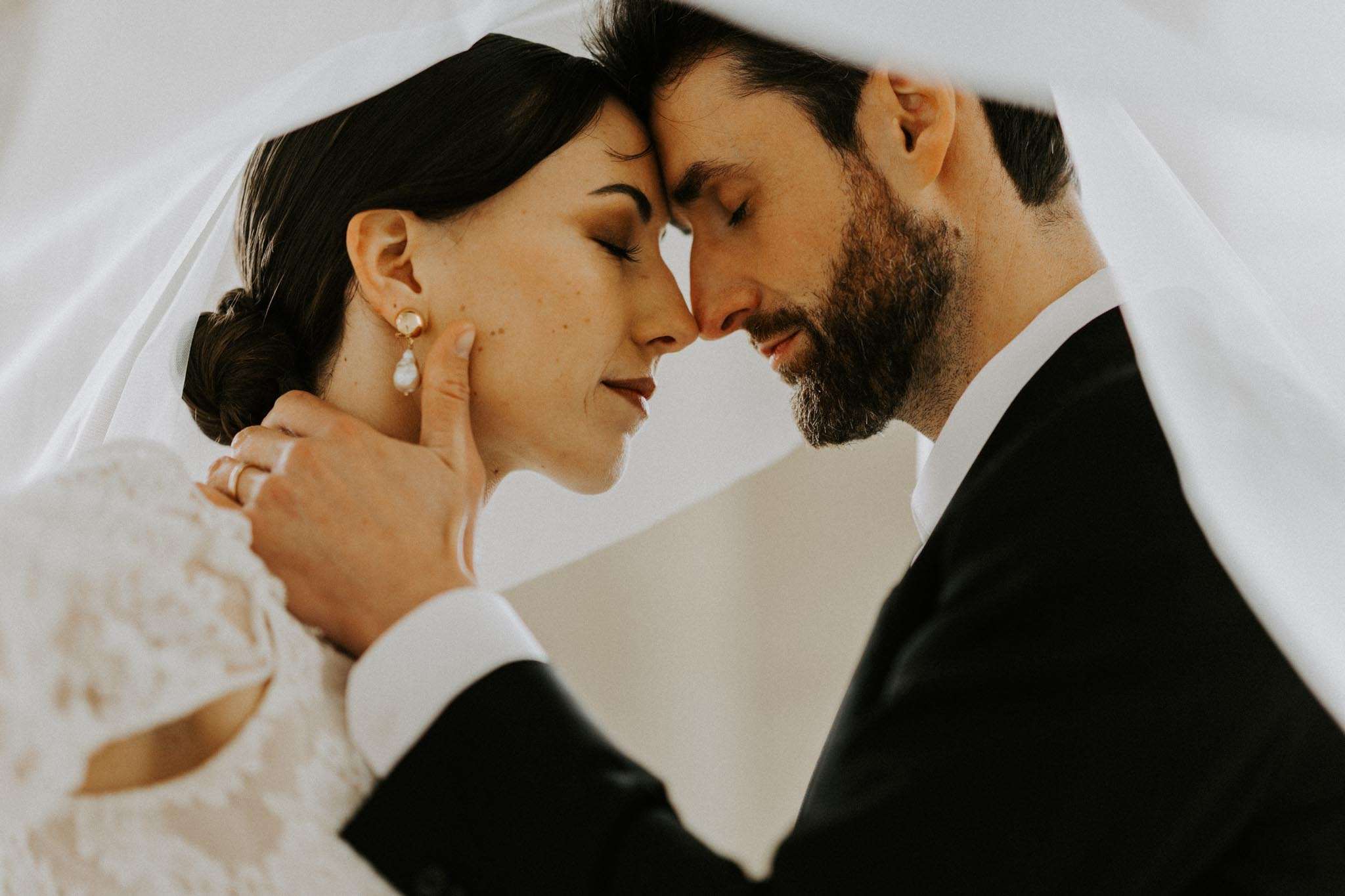 Bride and groom with foreheads touching under veil, bride in lace gown with gold pearl earrings
