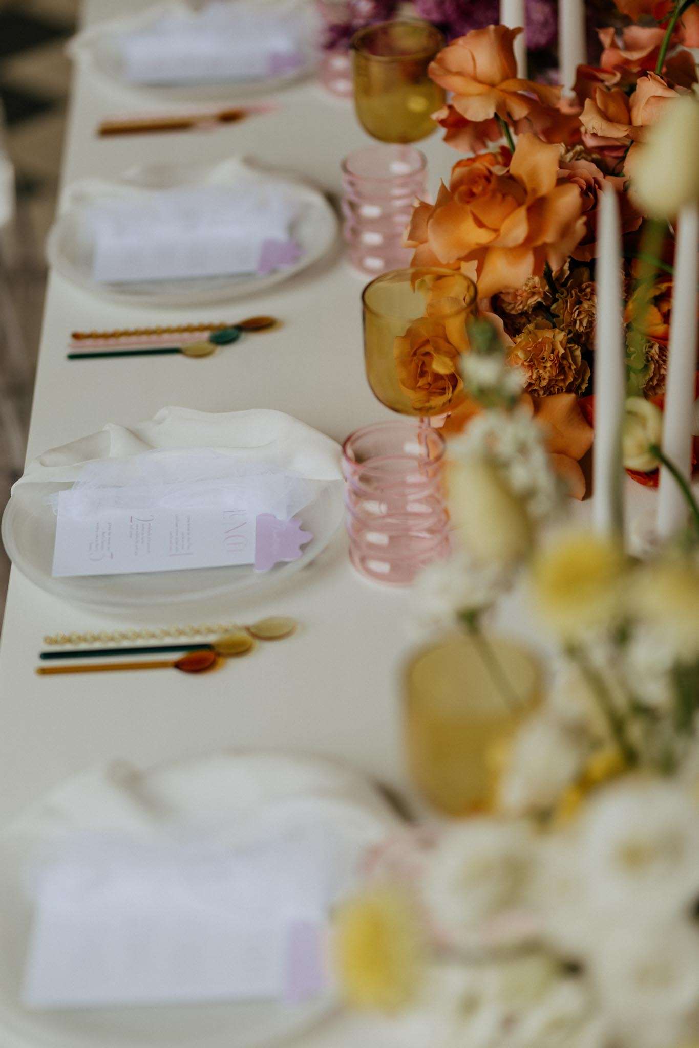 A close-up detail shot of a wedding reception tablescape set on a cream linen tablecloth. Each place setting features a white ceramic plate with a folded white linen napkin and a printed menu card with lavender accents, accompanied by gold and multicolored resin flatware in amber, green, and terracotta tones. The centerpiece runs along the length of the table and includes densely arranged orange and rust-toned garden roses, orange marigolds, and small white blooms, with tall white taper candles interspersed throughout. Colored glassware in ribbed pink and amber tones is placed at each setting, adding to the warm, retro-modern color palette that combines amber, terracotta, blush, and cream.