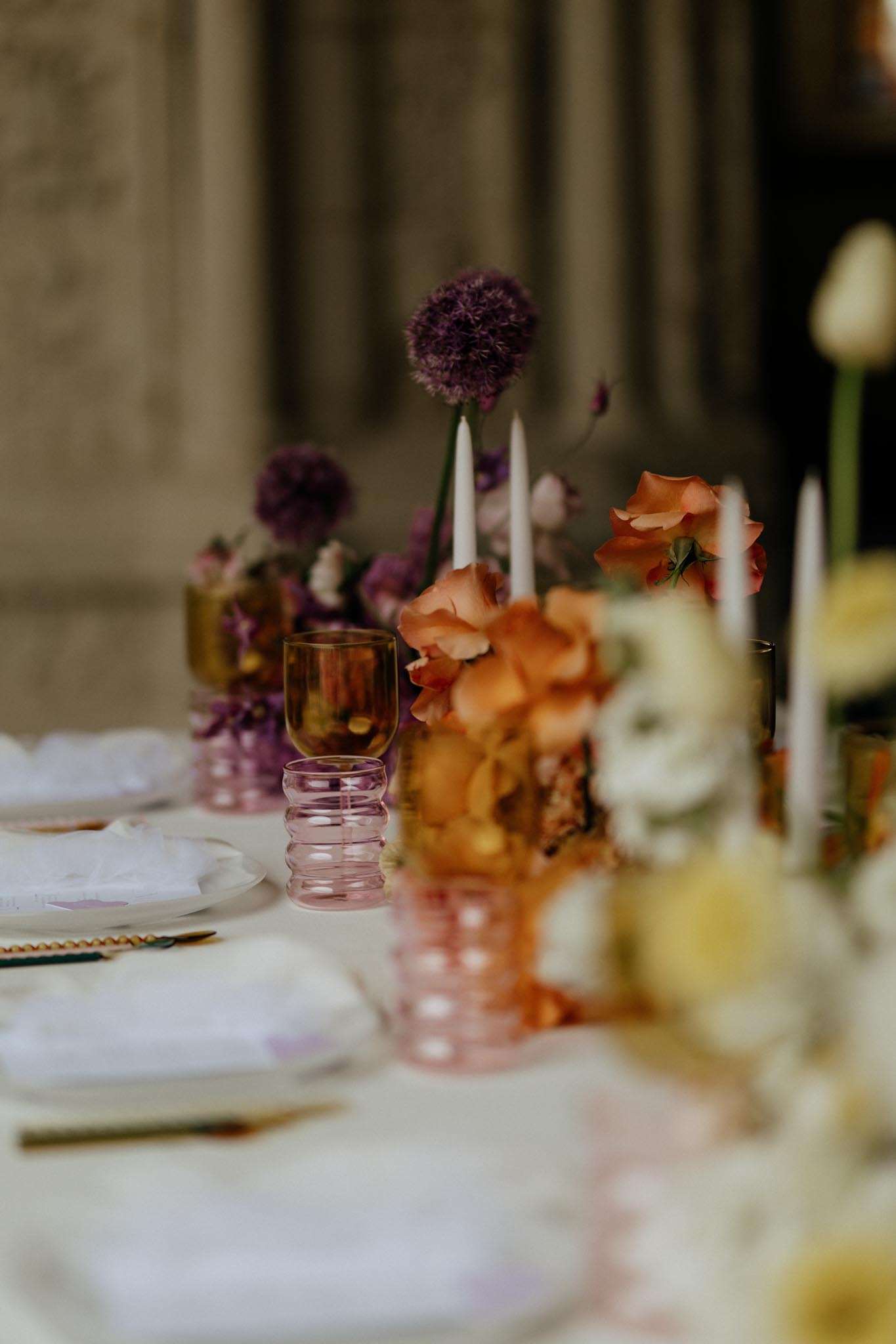 A close-up detail shot of a wedding reception tablescape styled in a rich, jewel-toned palette. The centerpiece arrangement features purple allium globe flowers, orange sweet peas, and cream ranunculus displayed in amber and pink bubble-glass vases, accompanied by slender white taper candles in matching colored holders. Place settings include white plates with folded white linen napkins, gold flatware, and a mix of amber and dusty pink ribbed glassware. The overall styling is modern with a bold, eclectic color palette combining terracotta, mauve, amber, and violet tones across both the florals and tableware.