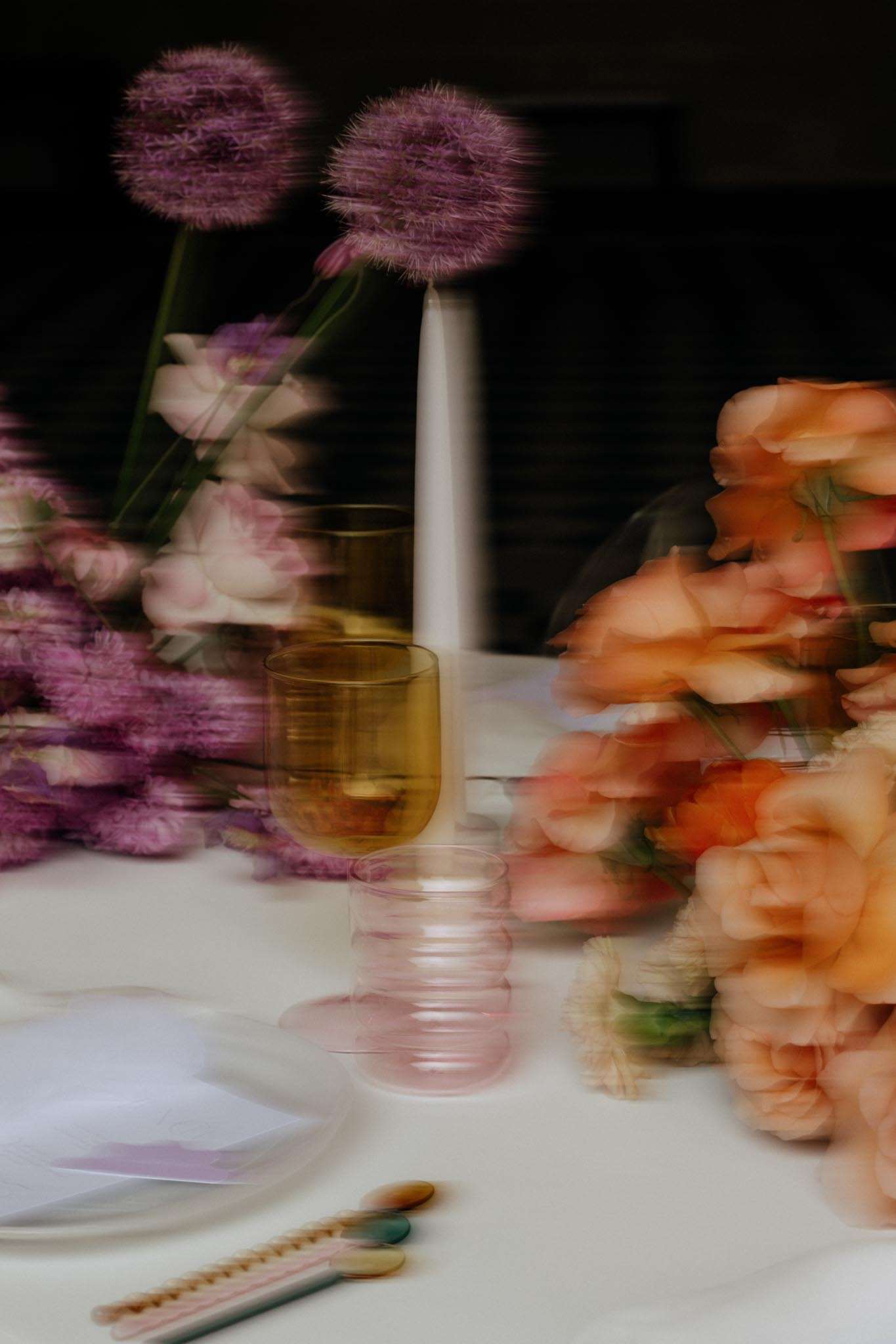 Motion-blurred reception tablescape with peach and coral roses, purple alliums, amber glassware, and taper candles