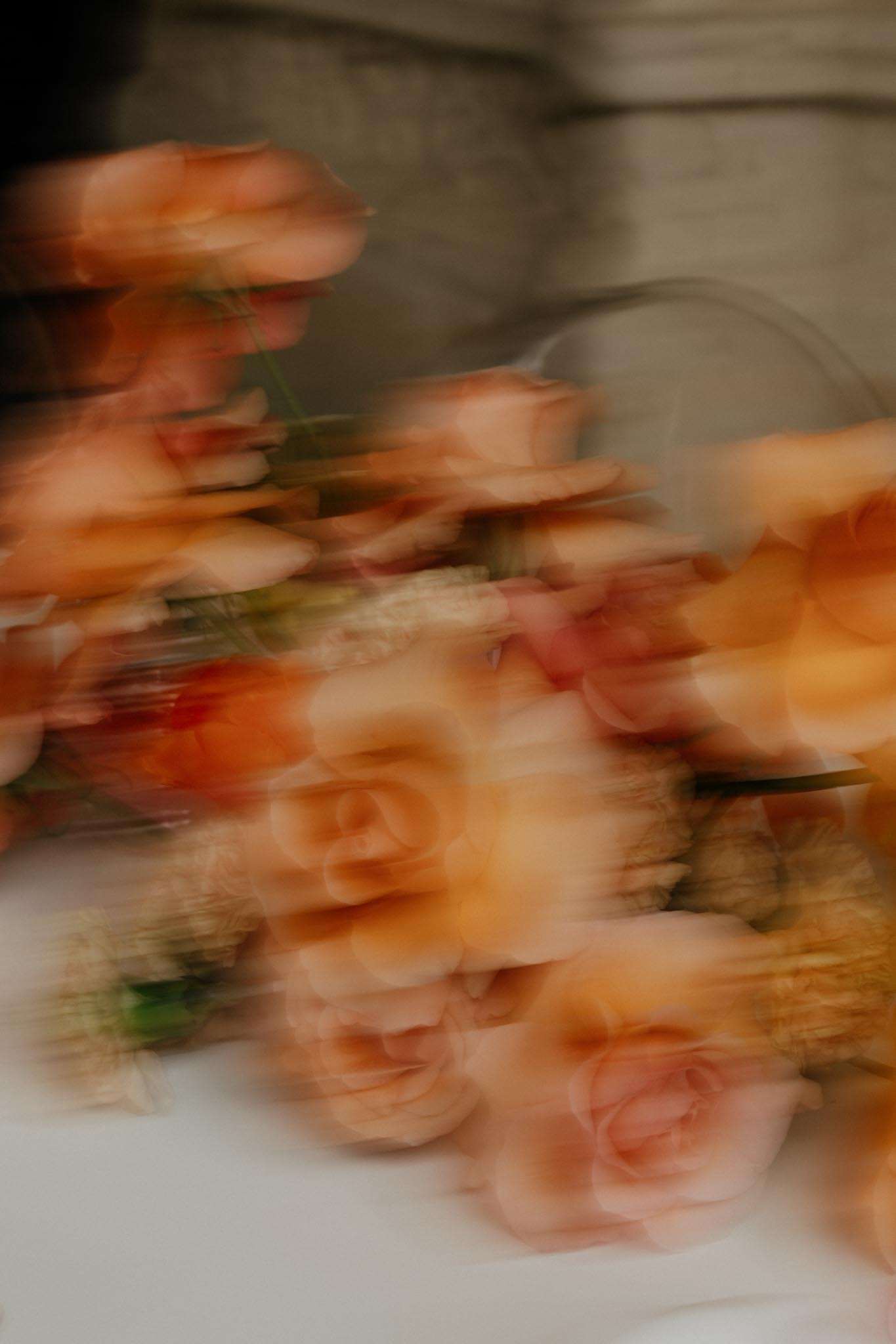 This image is heavily motion-blurred, likely an intentional creative technique. It appears to show a floral arrangement or bouquet featuring peach and apricot-toned roses mixed with what appears to be dried or textural elements in warm golden tones, resting on a white surface. The motion blur creates a layered, smeared effect across the entire frame, making individual blooms difficult to isolate. A stone architectural element is partially visible in the background.