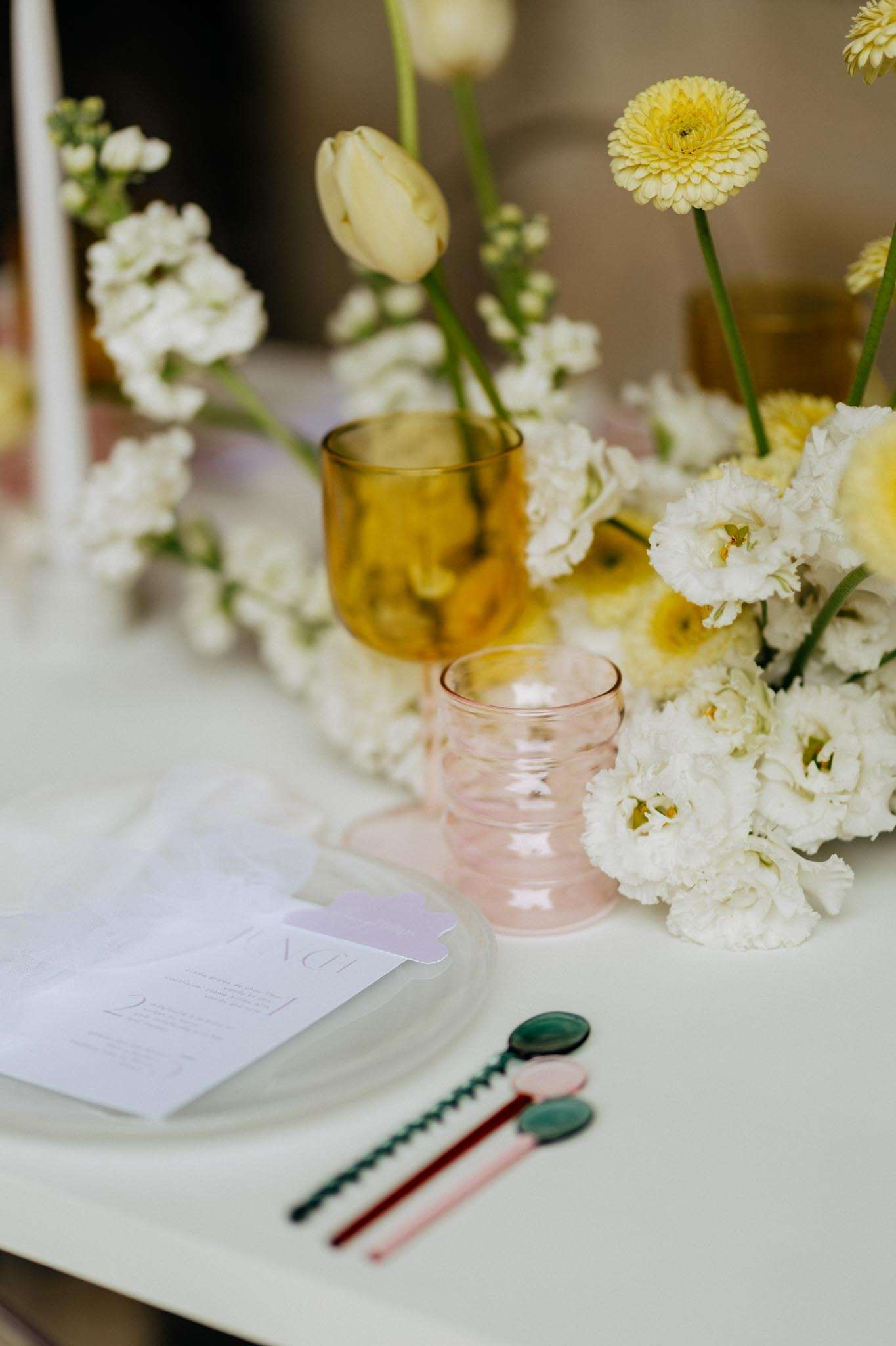 A close-up detail shot of a wedding reception table setting on a white linen tablecloth. The centerpiece features a low, loose arrangement of white lisianthus, white stock flowers, cream tulips, and yellow gerbera daisies with tall green stems. Two colored glass goblets are placed among the florals — one amber/mustard-toned and one blush pink with a ribbed texture. At the place setting, a white plate holds a printed menu card in pale lavender and white tones. Two decorative spoons with teal green oval handles and twisted red and pink handles are laid beside the plate, adding a playful, modern touch. The overall palette combines white, cream, yellow, blush, and amber with a contemporary, color-forward styling approach.