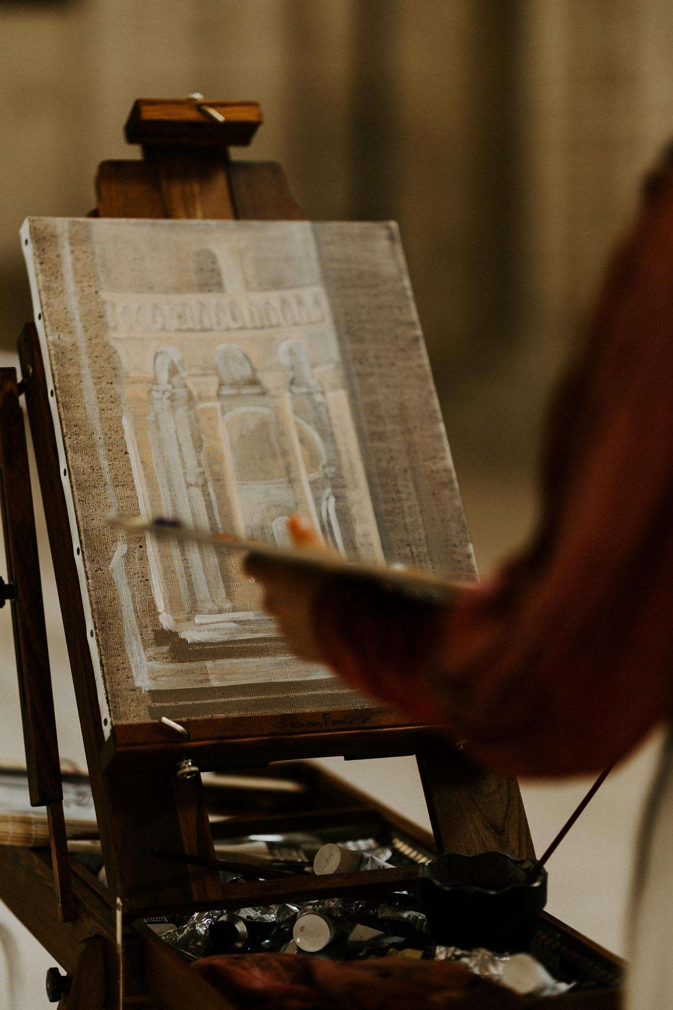 Close-up of a live wedding painter's hand adding brushstrokes to an oil painting of a venue facade on an easel