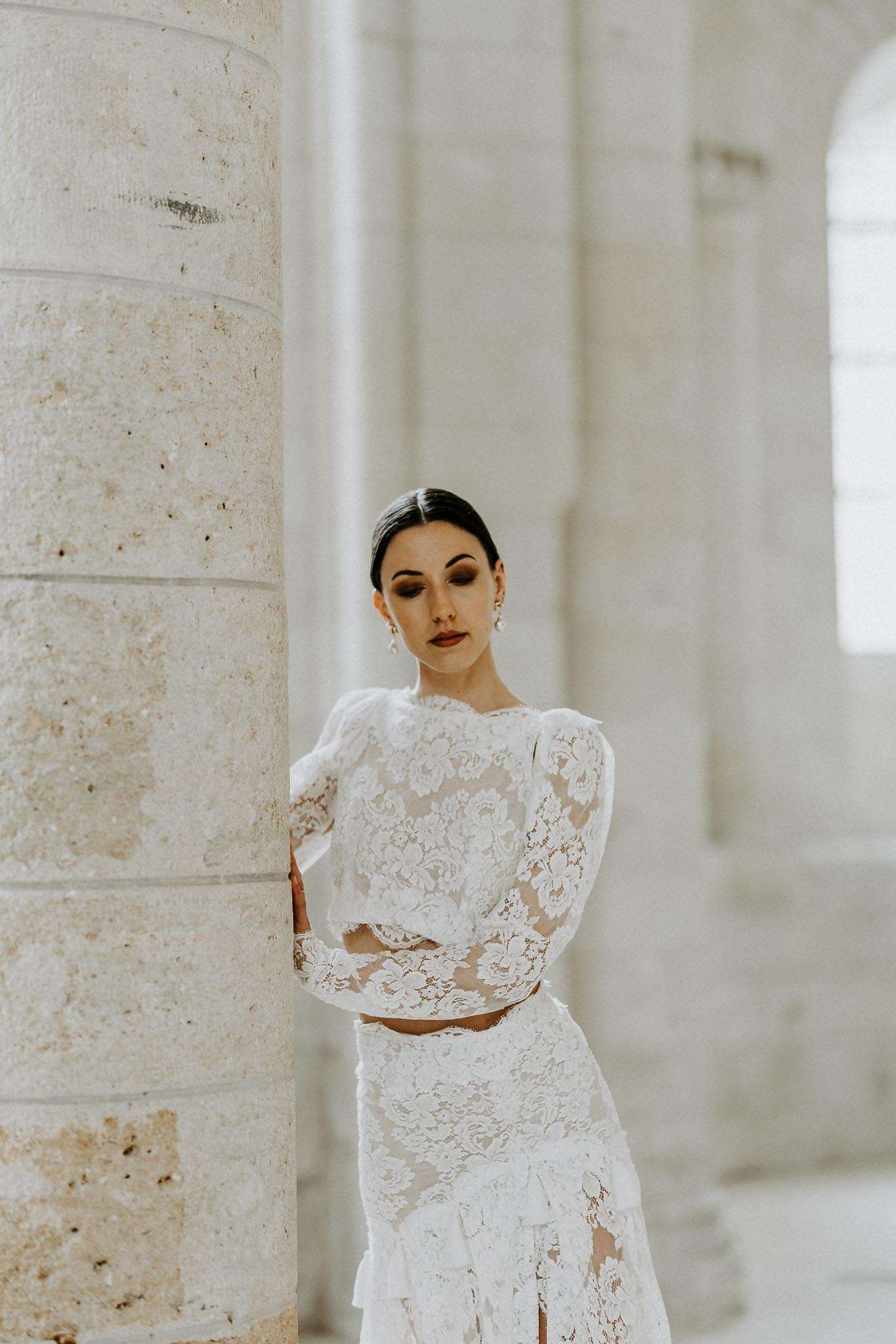 A bridal portrait of a single bride posing beside a large cylindrical stone column inside what appears to be a historic chapel or abbey interior, with arched windows and pale stone architecture visible and softly blurred in the background. The bride wears a two-piece white lace bridal ensemble consisting of a long-sleeved crop top with puffed shoulders and floral lace pattern, paired with a matching fitted lace skirt featuring tiered ruffle detailing and a slight slit. Her dark hair is pulled back into a sleek low bun, and she wears drop pearl earrings with bold eye makeup in warm brown tones. The composition is a close-up portrait shot with shallow depth of field, with the column occupying the left third of the frame and the bride gazing downward.