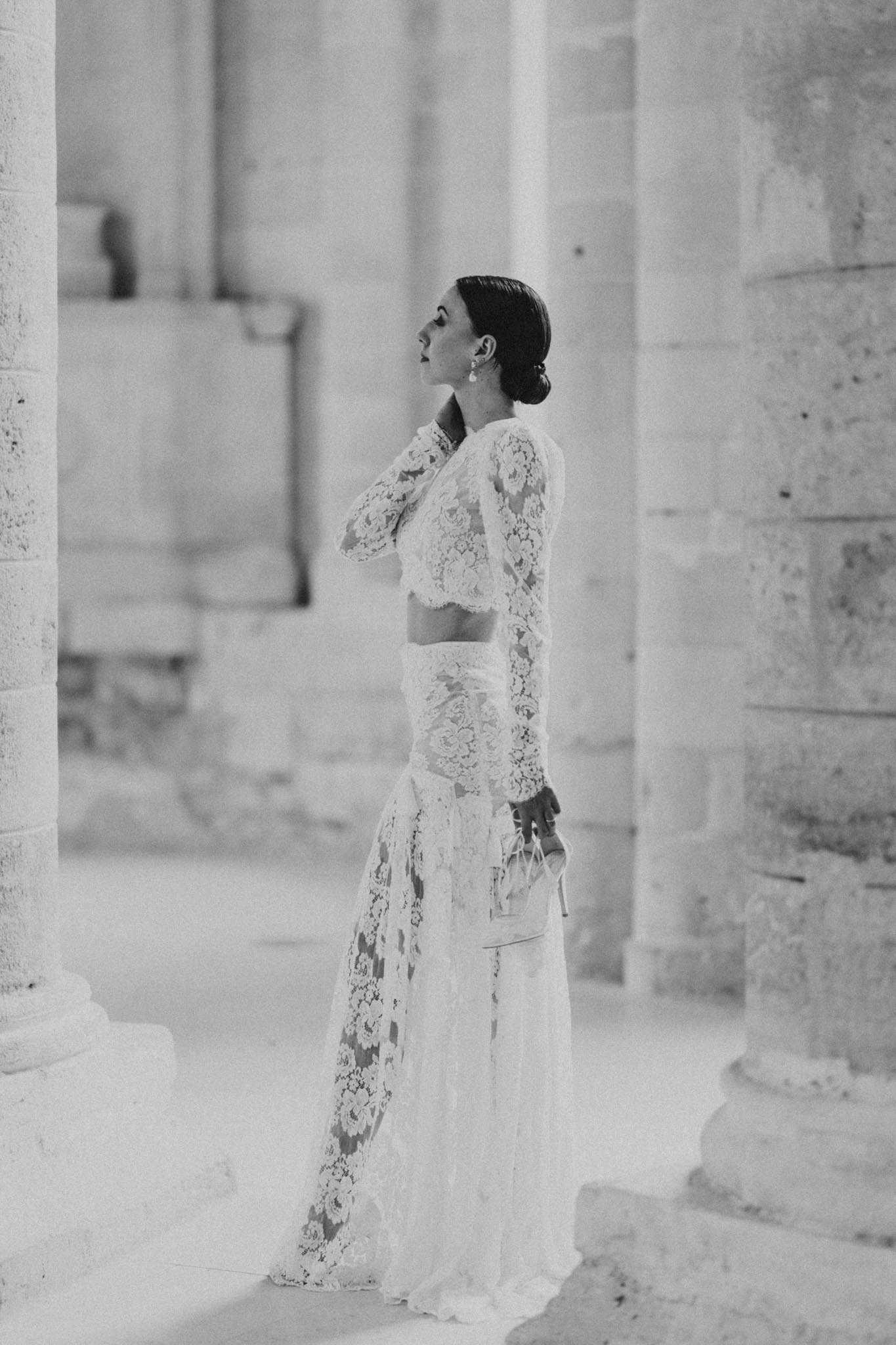 Bride in two-piece lace ensemble standing between stone columns gazing upward in high-key B&W