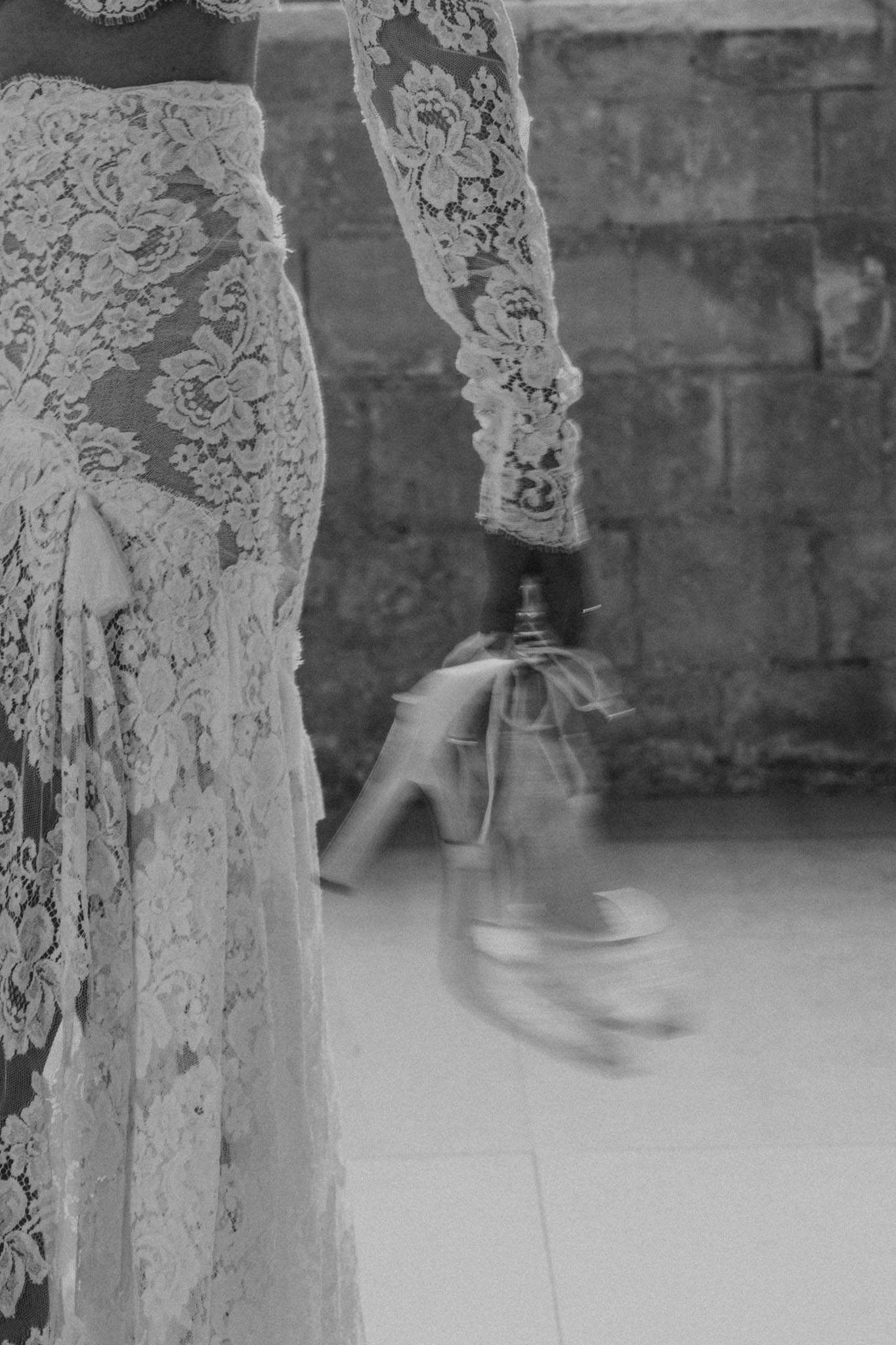A black-and-white close-up detail shot of a bride during what appears to be a getting-ready moment, set against a rough stone wall background typical of a French château or historic venue. The image focuses on the lower torso and arm of the bride, showcasing a fitted long-sleeve lace wedding dress with intricate floral lace patterns, a scalloped lace cuff, and a layered lace skirt with a small fabric bow detail at the hip. The bride's hand holds a pair of heeled bridal shoes with ribbon tie detailing, which appear slightly motion-blurred, adding a sense of movement to the composition. The high-contrast black-and-white tones emphasize the texture of the lace against the light background.