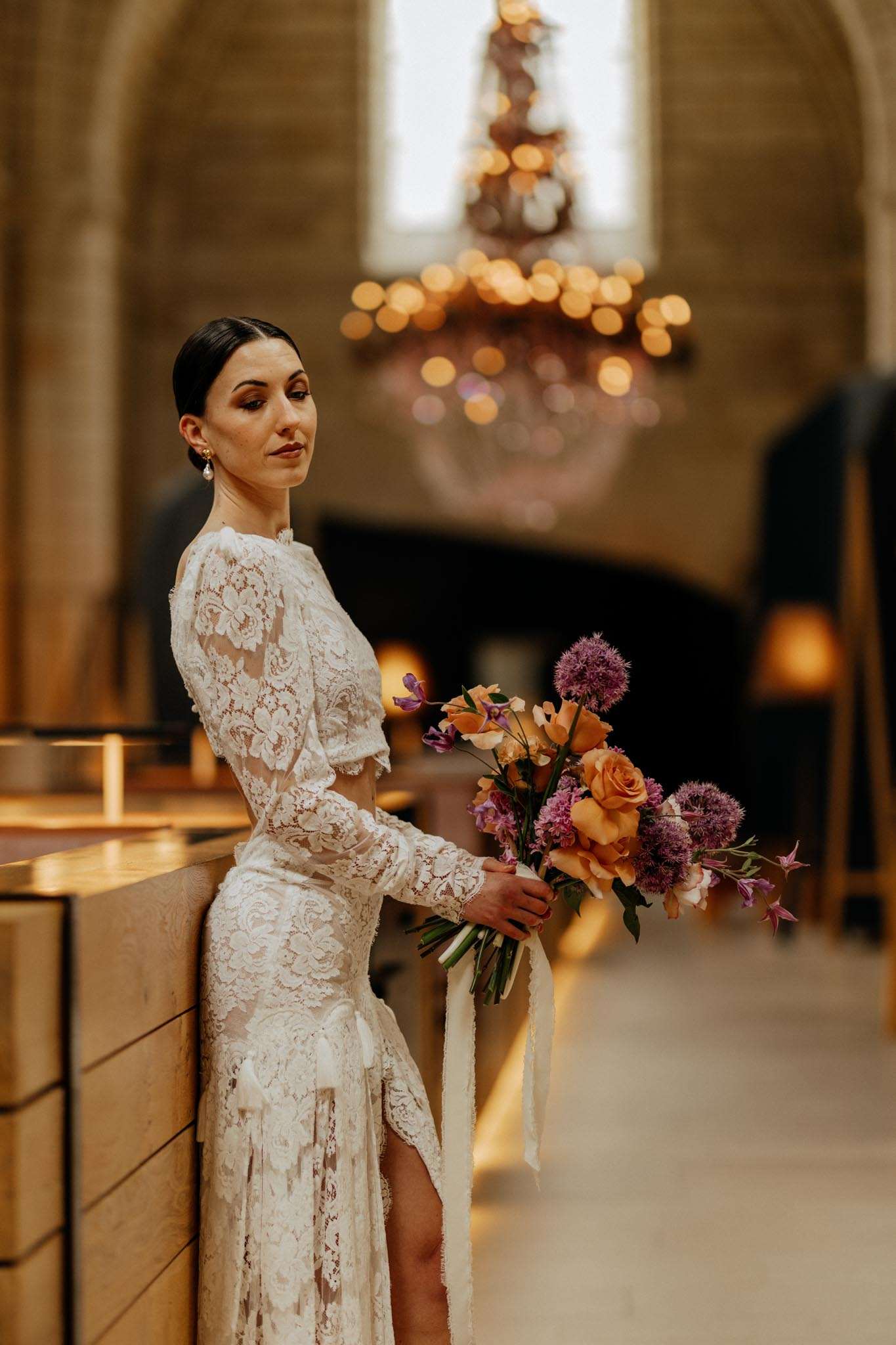 Bride in ivory lace two-piece outfit holding garden bouquet of peach roses and purple allium in candlelit chapel
