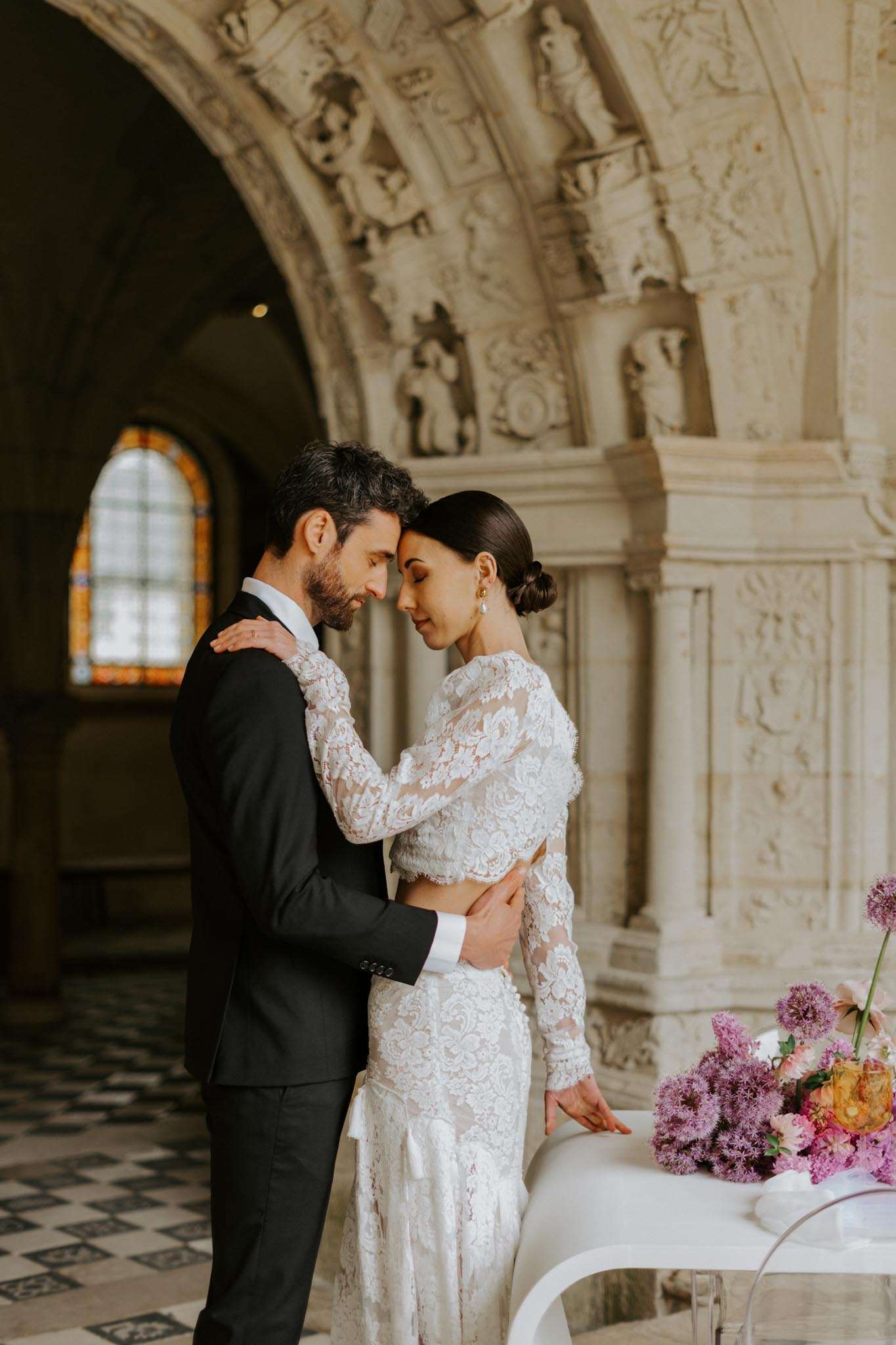 A couple portrait taken indoors inside a historic chapel or château with heavily carved cream stone archways, ornate sculptural relief detailing, and a stained glass window visible in the background. The bride wears a long-sleeve, fitted lace gown with a nude/ivory lace overlay and has her dark hair styled in a sleek low bun with pearl drop earrings; the groom wears a dark navy or black suit with a white shirt. They stand forehead-to-forehead in a close embrace, eyes closed. In the foreground to the right, a white table with a clear acrylic chair holds a floral arrangement featuring purple alliums, lilac ageratum, and orange and pink blooms. The floor is laid with a black and white geometric tile pattern. The overall styling is modern classic with bold, colorful florals contrasting against the pale stone architecture. Medium portrait shot.