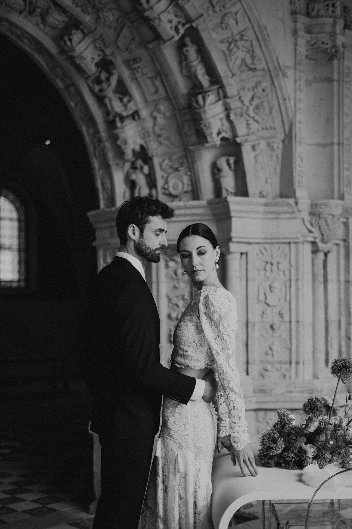 This is a black-and-white couple portrait taken indoors inside a grand historic building, likely a chapel or ornate château interior, featuring heavily carved stone archways with figurative relief sculptures and arched windows visible in the background. The groom, wearing a dark suit, stands closely behind the bride, his hand resting at her waist, both looking downward in a quiet, still pose. The bride wears a long-sleeve fitted lace gown with an intricate floral lace pattern across the bodice and sleeves, her hair pulled back in a sleek low bun, and she wears drop earrings. To the right of the frame, a partially visible floral arrangement sits on what appears to be a white surface, featuring large rounded blooms and tall stemmed flowers with a loose, sculptural composition. The image uses deep shadow and soft mid-tones with strong contrast, and the architectural backdrop gives the shot a formal, classical feel. The composition is a medium portrait shot framing the couple from roughly the waist up.