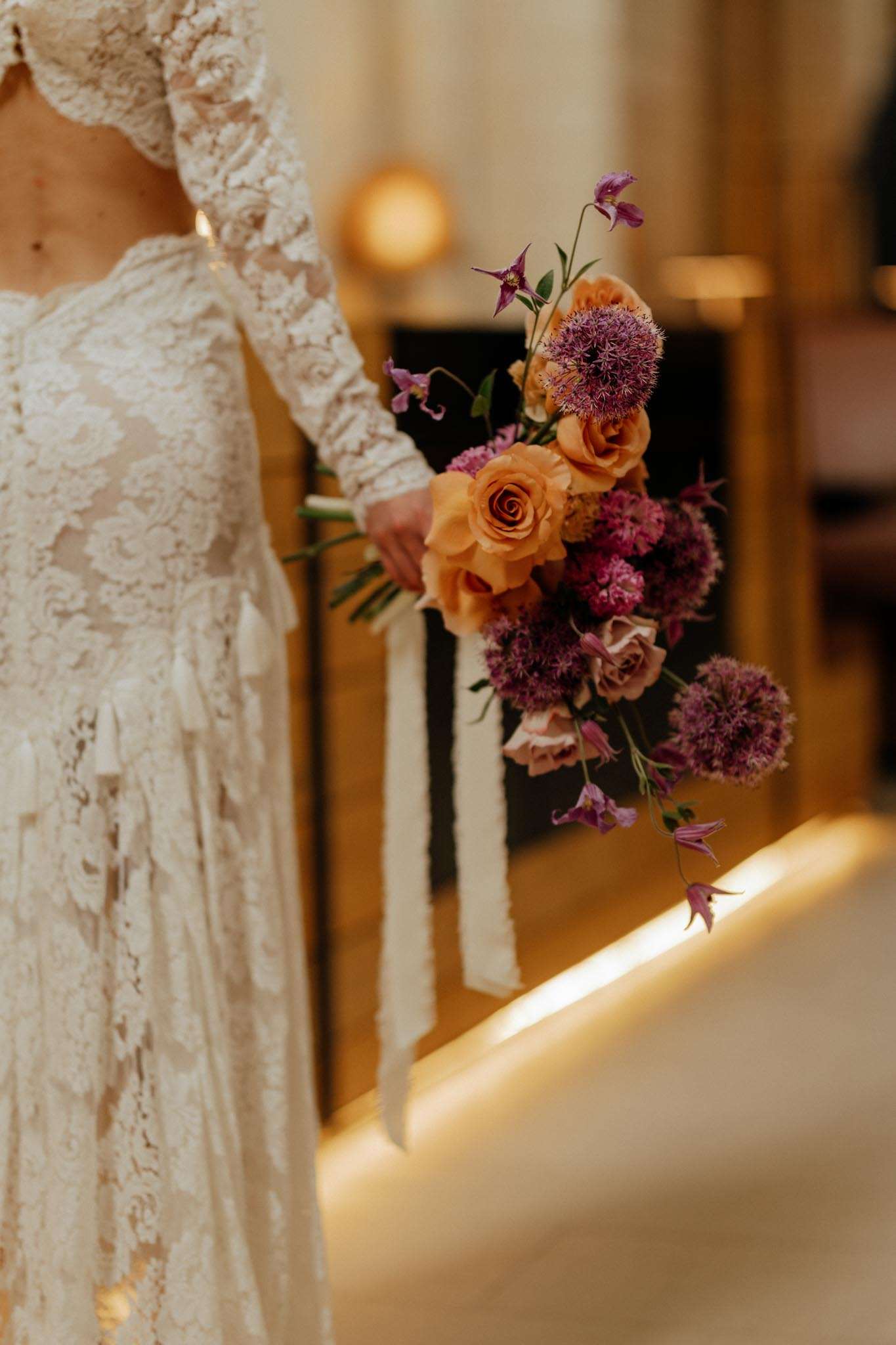 Bride in long-sleeve lace gown holding garden-style bouquet of terracotta roses, purple allium, and mauve blooms