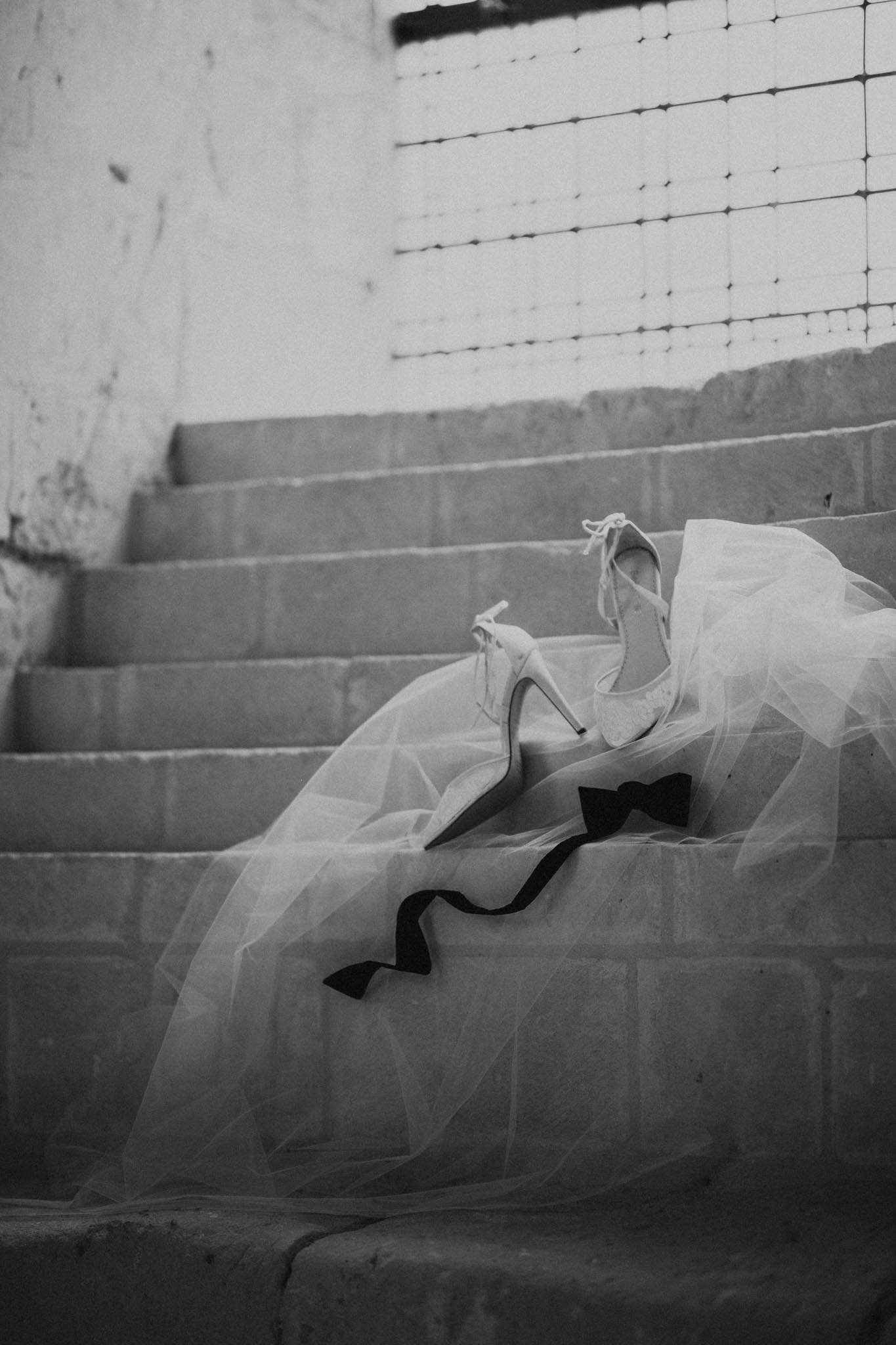 A black-and-white detail shot of bridal accessories arranged on wide stone steps, likely inside a venue with a tiled or glass-block window visible in the background. A pair of white stiletto heels with ankle-tie straps and lace detailing rest on top of a flowing tulle veil or skirt, which cascades across several steps. A dark black bow tie is placed alongside the shoes on the tulle fabric, pairing bridal and groom accessories together in a single styled composition. The image has soft, diffused light with mid-range contrast and no people present.