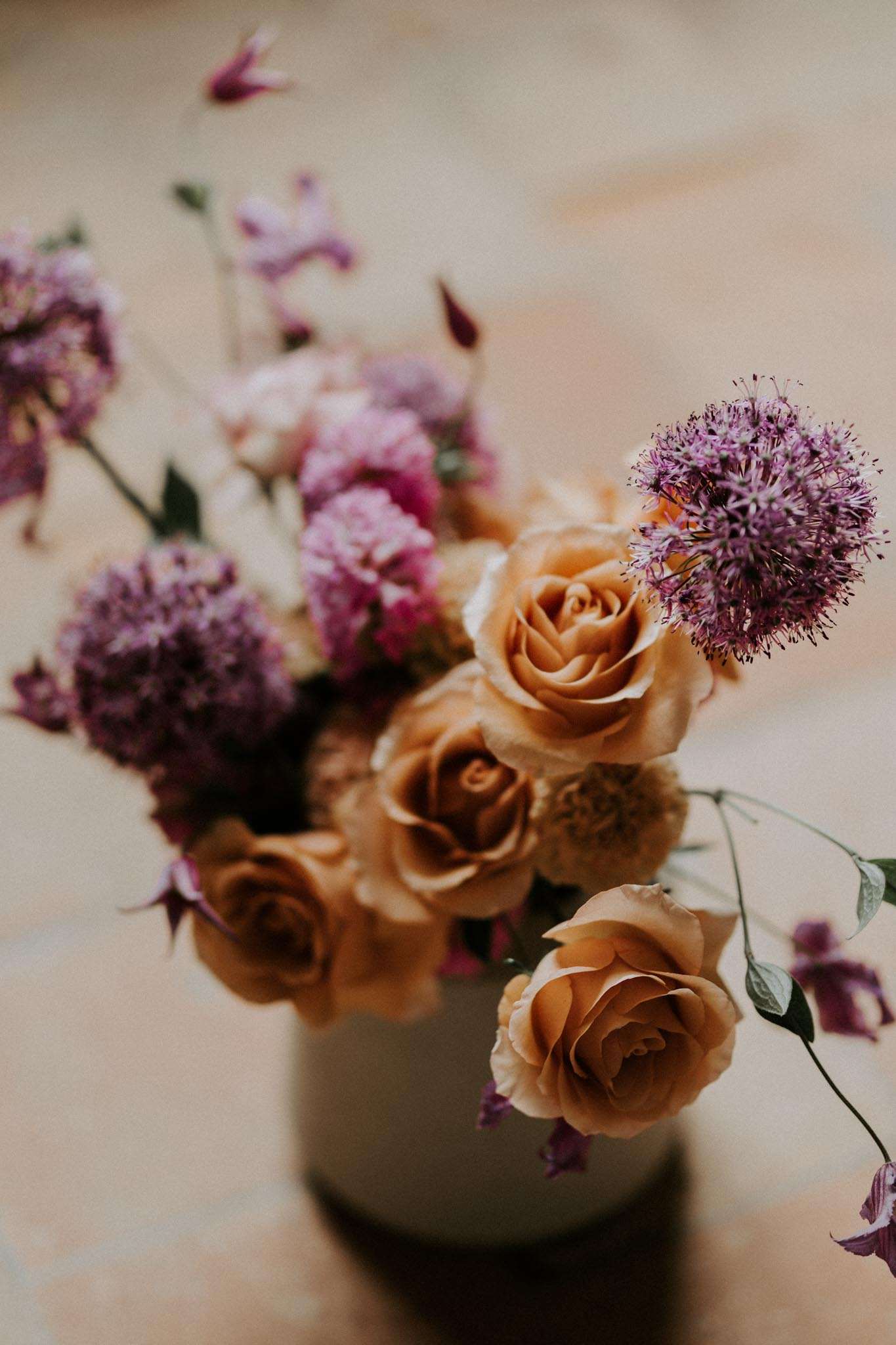 Close-up detail shot of a floral arrangement in a small matte grey ceramic vase, placed on a flat surface with a soft blurred neutral background. The arrangement features tawny amber-orange garden roses as the focal blooms, combined with purple allium spheres, mauve and magenta scabiosa, purple sweet peas or columbines, and delicate green trailing foliage. The color palette is warm terracotta and amber tones contrasted with cool violet and mauve purples, giving the arrangement a rich, garden-gathered aesthetic. The shallow depth of field keeps the foreground roses sharp while the background blooms fall into soft focus.