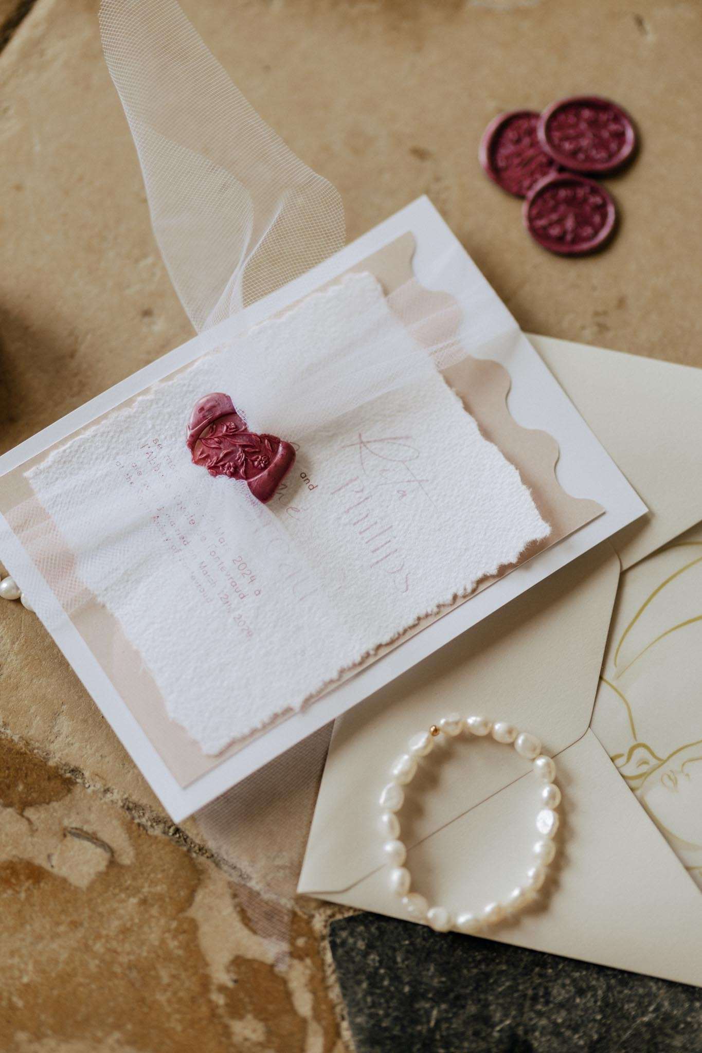Layered invitation suite with torn-edge paper, burgundy wax seals, and pearl bracelet on stone