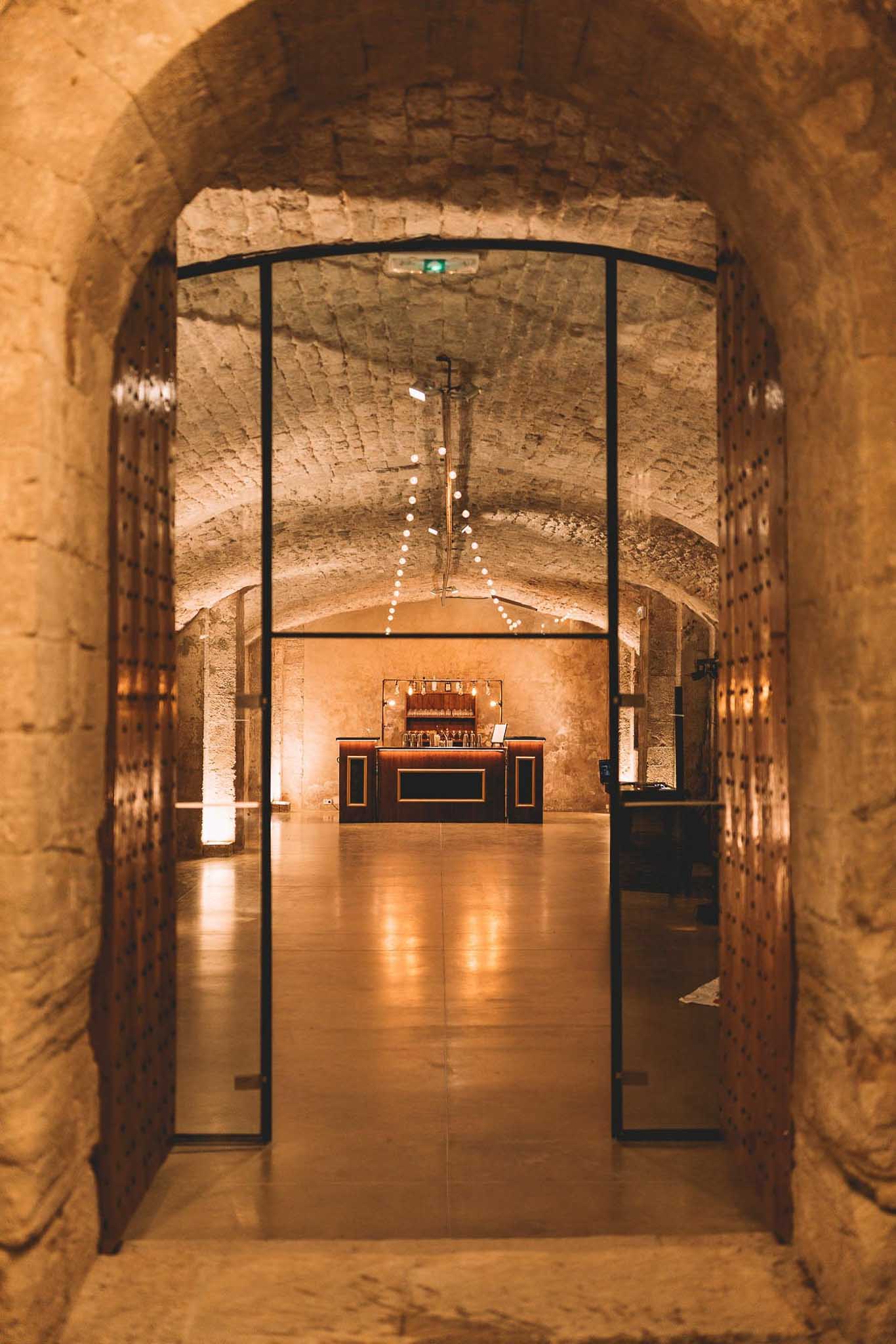 Vaulted stone cellar bar room with amber uplighting and globe string lights viewed through a glass-panelled archway