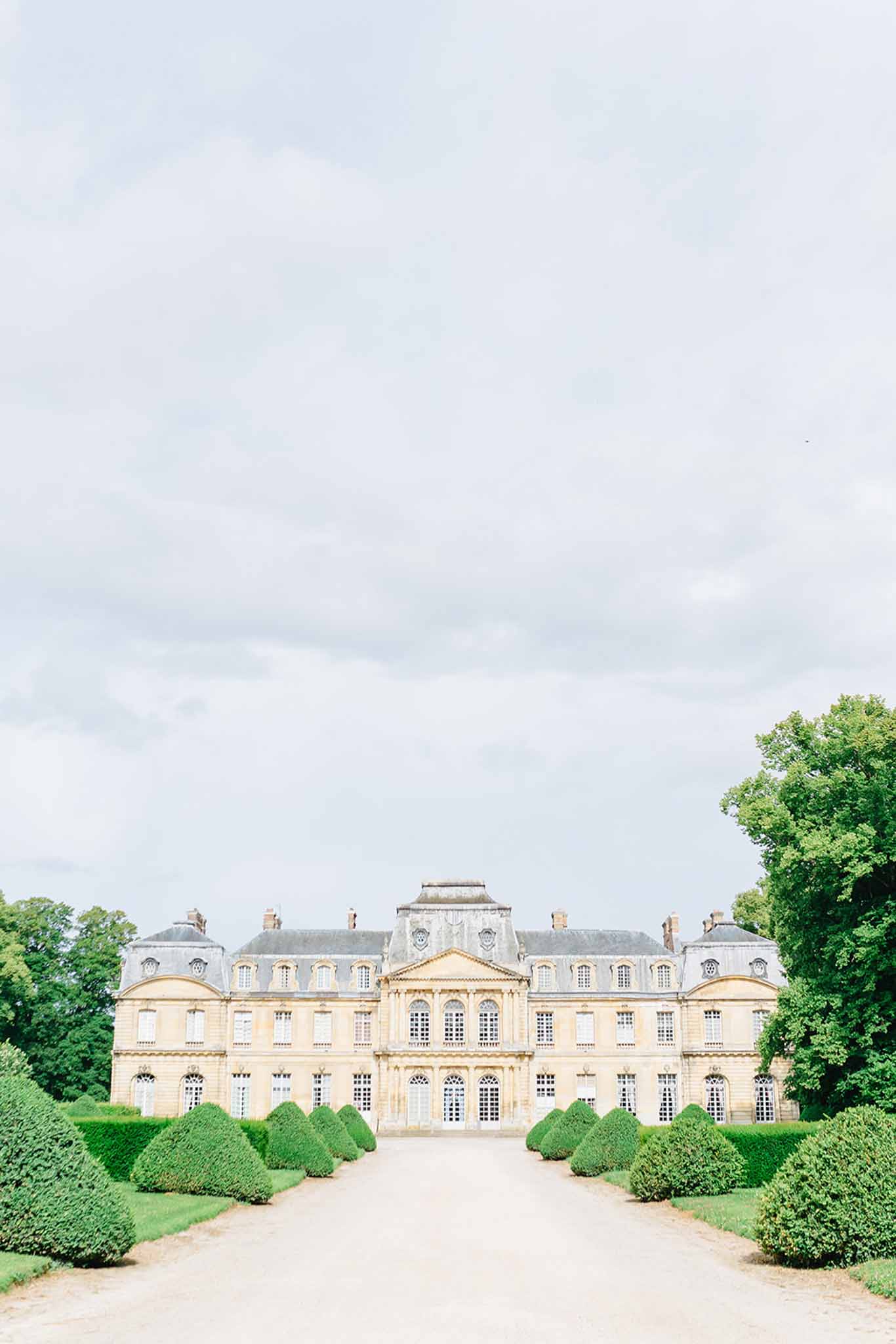 Grand French château façade with symmetrical mansard roofs and clipped boxwood topiaries lining gravel approach