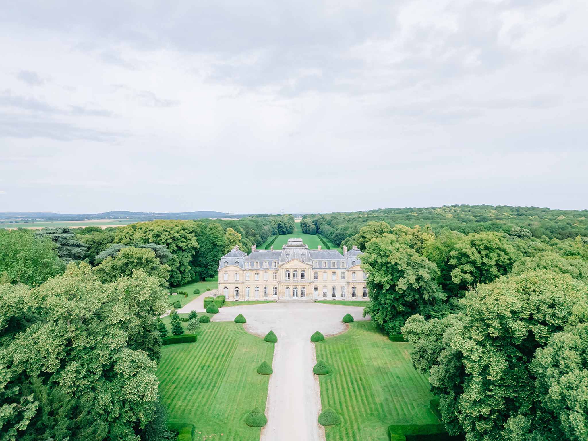 Aerial view of classical French château with mansard roof and symmetrical architecture flanked by cone topiaries and dense woodland