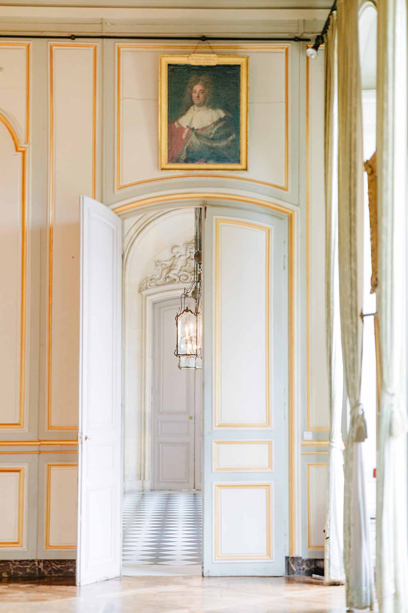 Classical interior gallery with cream and gold paneling, gilded portrait above arched doorway and curtained windows