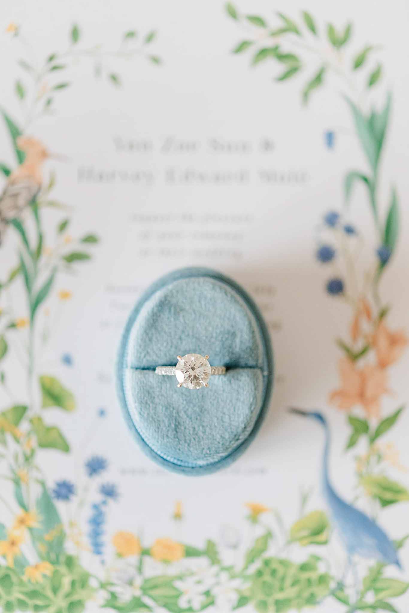 Diamond engagement ring in dusty blue velvet box positioned on botanical watercolor wedding stationery