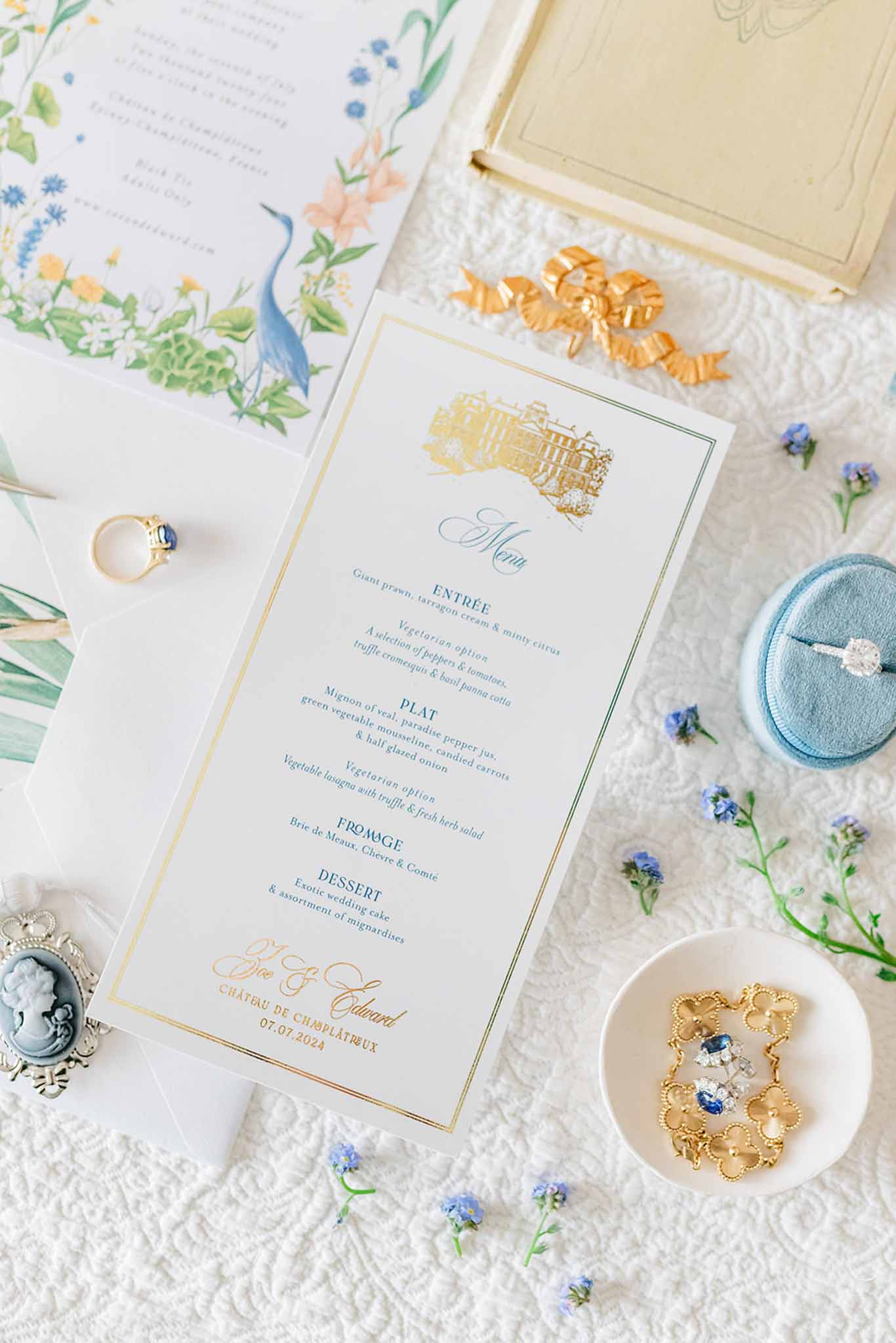 Flat lay of wedding stationery with gold borders, sapphire ring, blue forget-me-nots, and watercolor botanical cards