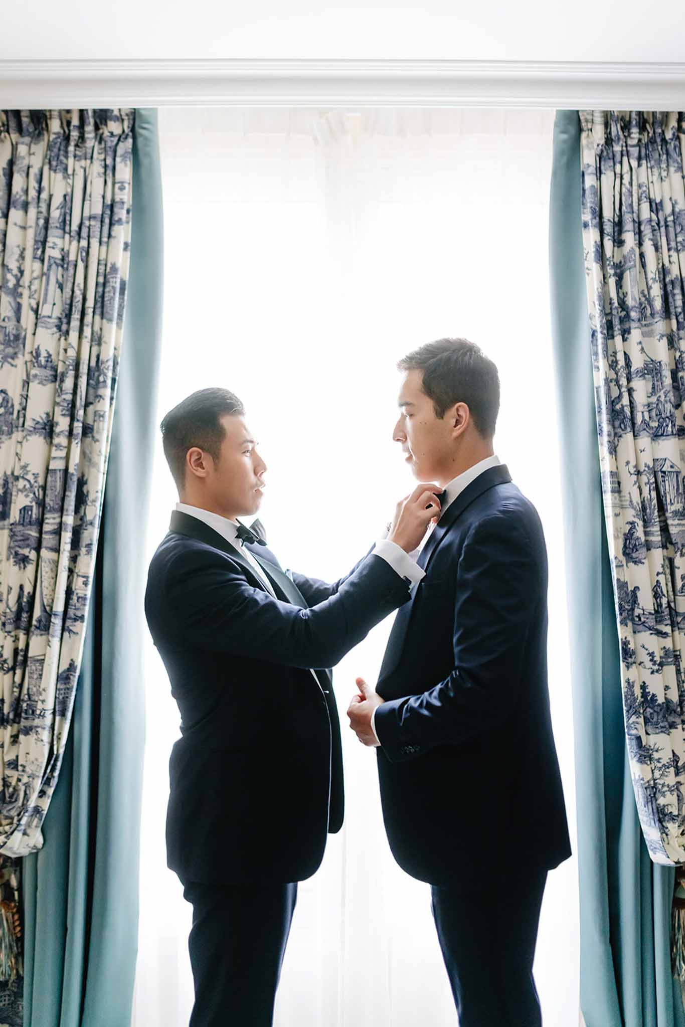 Two grooms adjusting bow tie in room with Toile de Jouy curtains during getting-ready preparations