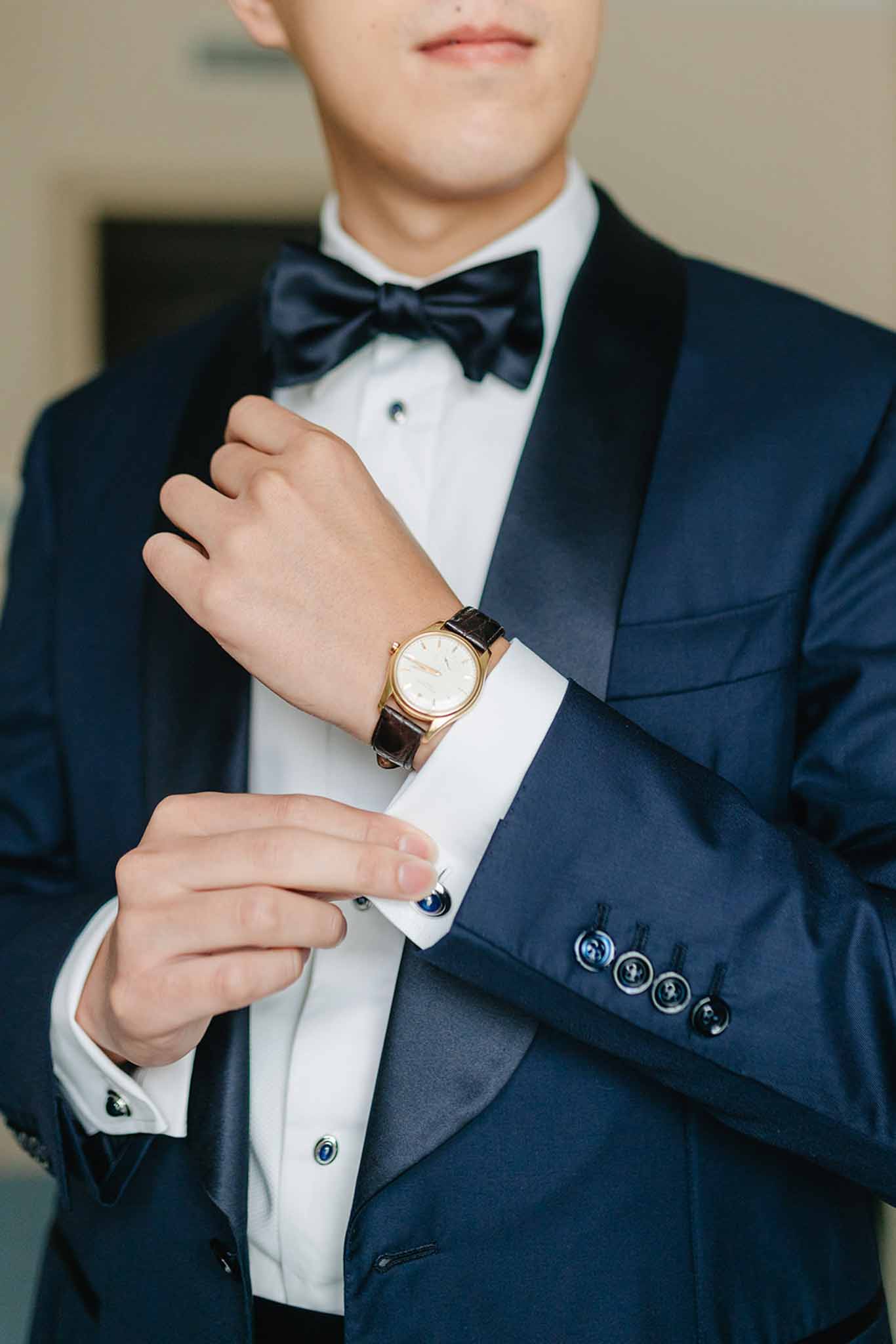 Close-up of groom in navy tuxedo showing white shirt, navy bow tie, gold watch, and blue stone cufflinks