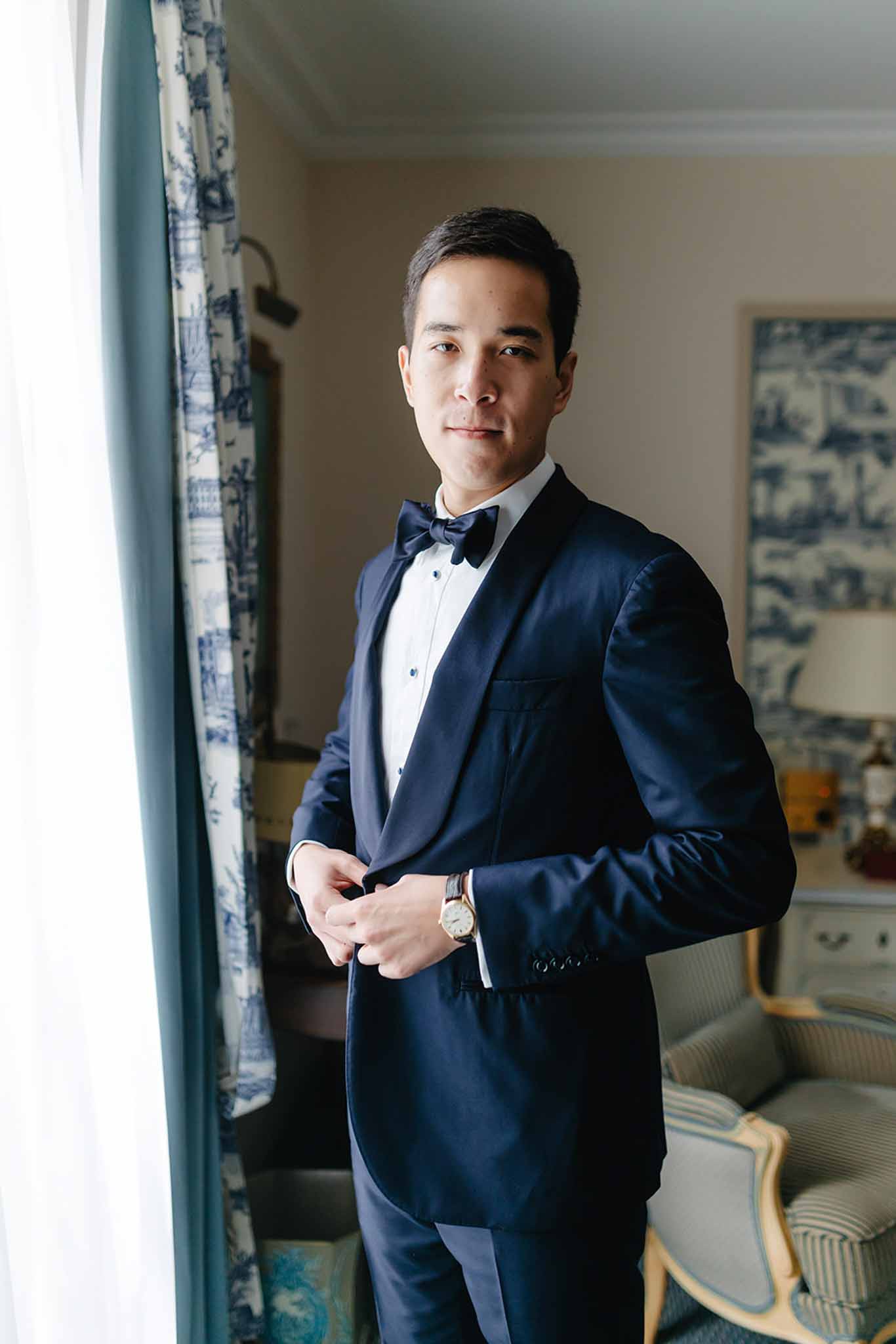 Groom in navy tuxedo and bow tie standing by window during getting ready preparations in classic hotel room