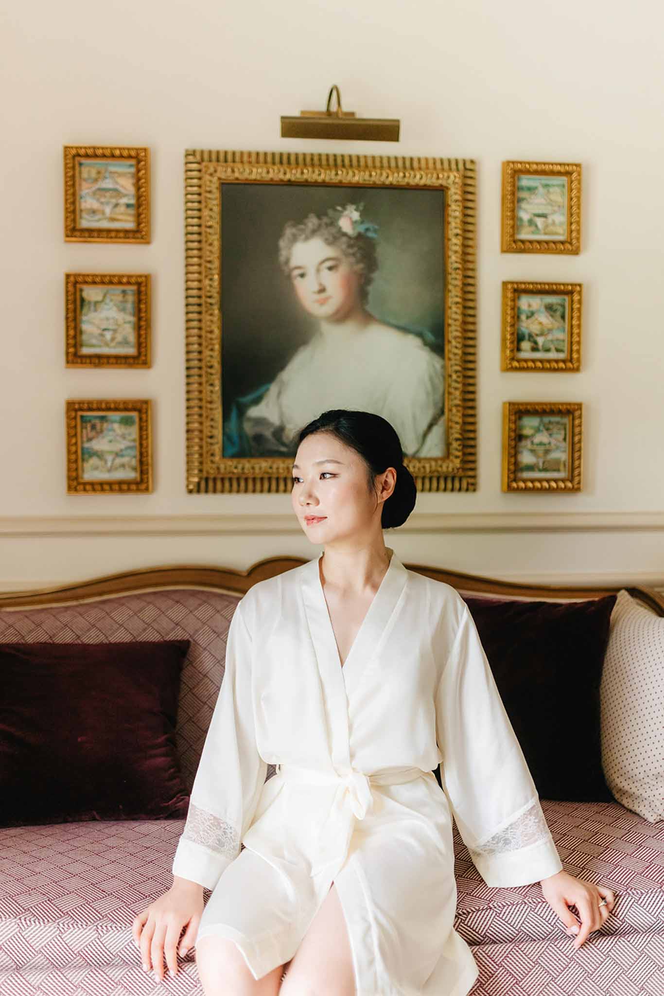 Bride in cream silk robe sits on patterned sofa in ornate chateau room with gold-framed paintings on the wall
