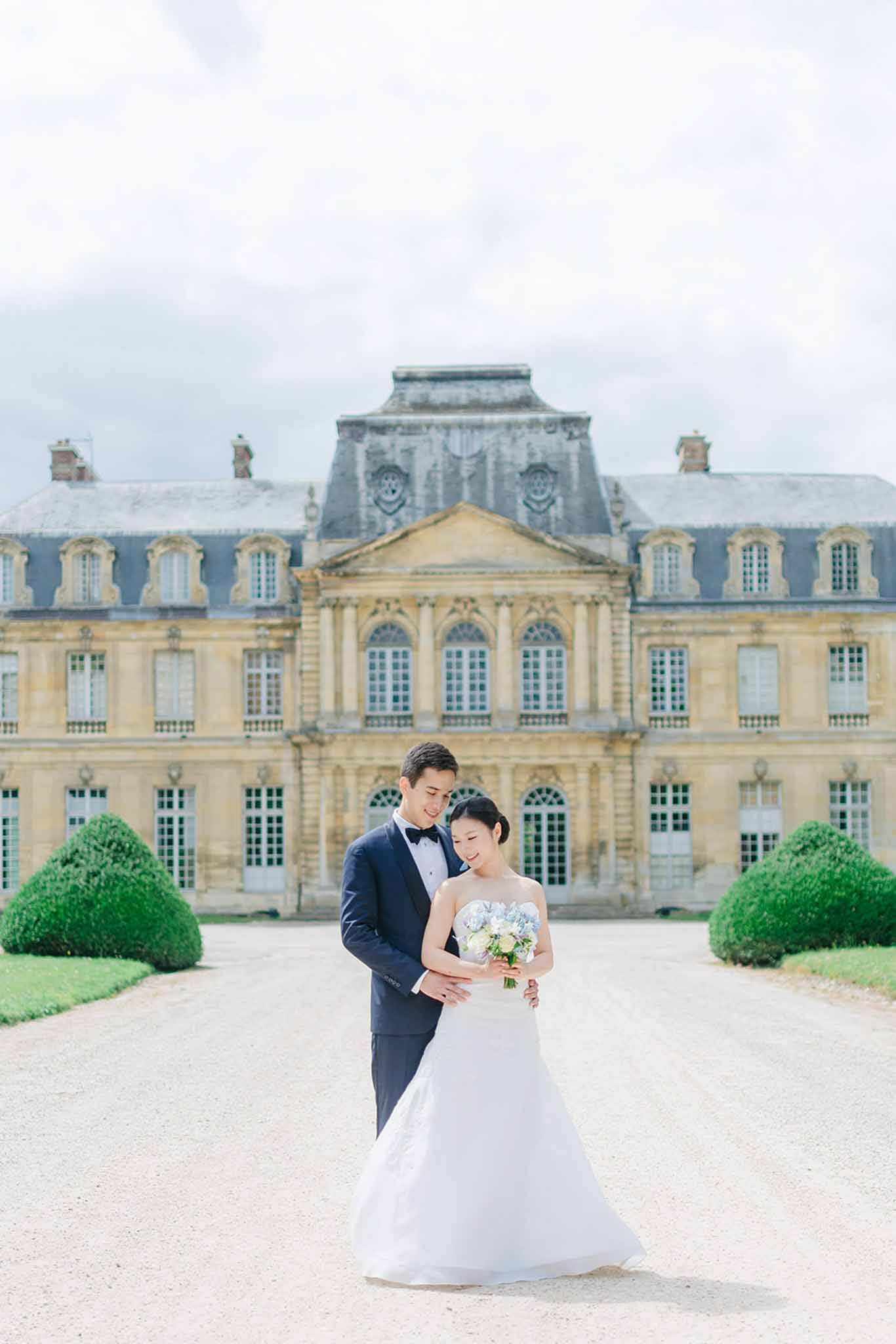 Bride and groom in close portrait embrace on gravel courtyard with classical limestone chateau behind them