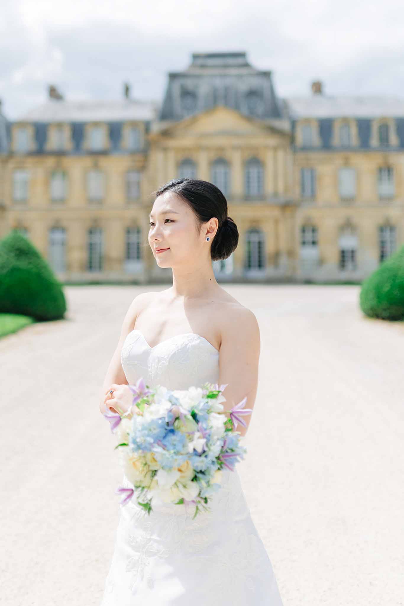 Bride in white lace gown holding bouquet of blue hydrangeas and peach roses before grand yellow stone palace