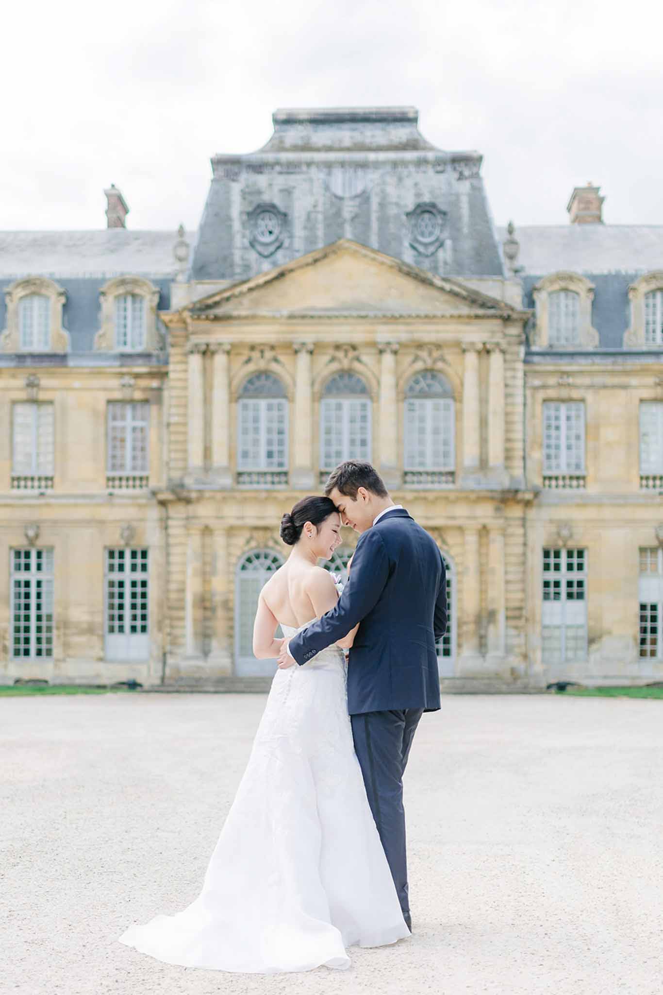 Bride and groom in intimate portrait with foreheads touching on chateau courtyard with mansard roofline behind