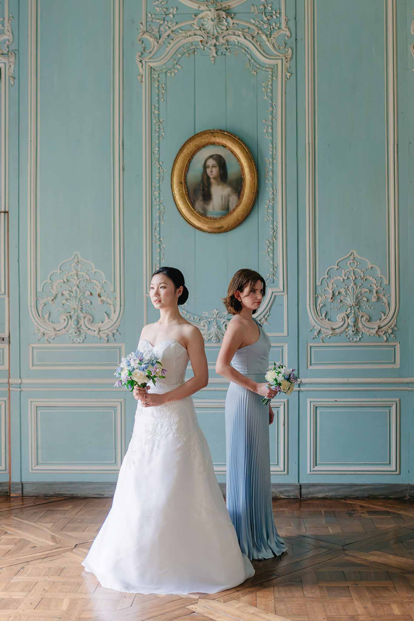 Bride in ivory beaded strapless gown beside bridesmaid in slate blue pleated dress in palatial turquoise-panelled room