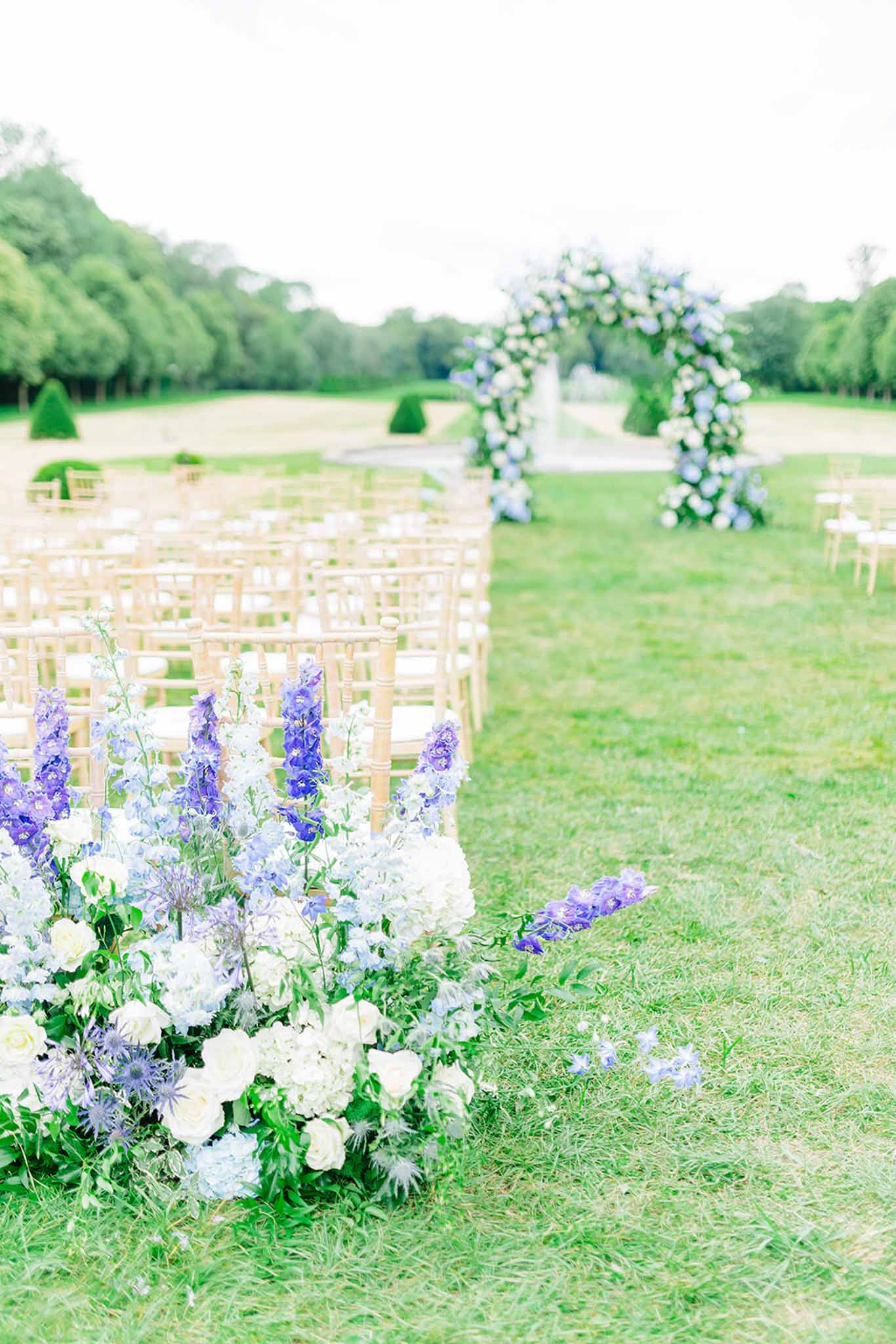 Outdoor ceremony aisle lined by chiavari chairs leading to floral arch of white and periwinkle blue hydrangeas