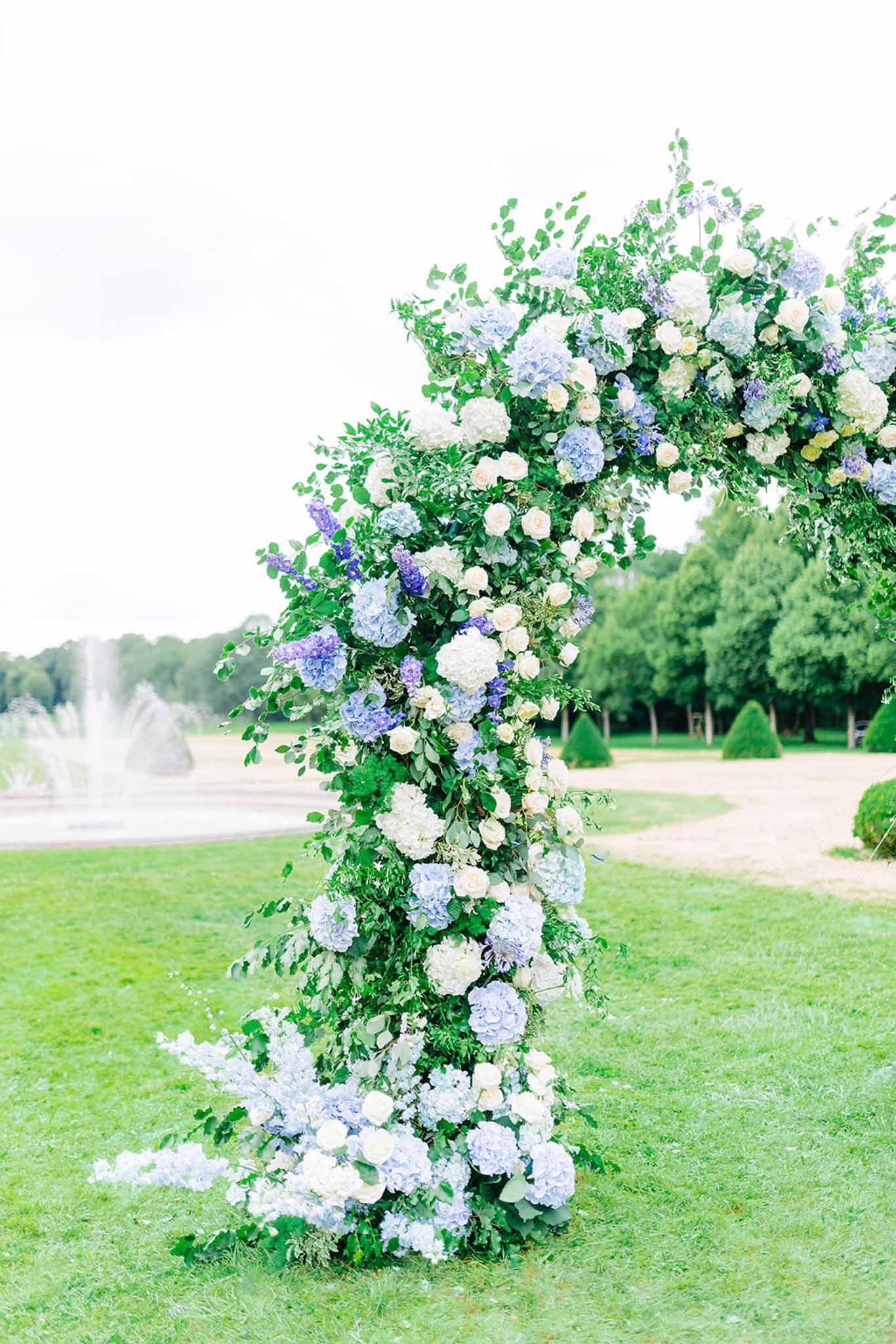 Tall floral arch of ivory and lavender hydrangeas, white roses and greenery in formal estate garden