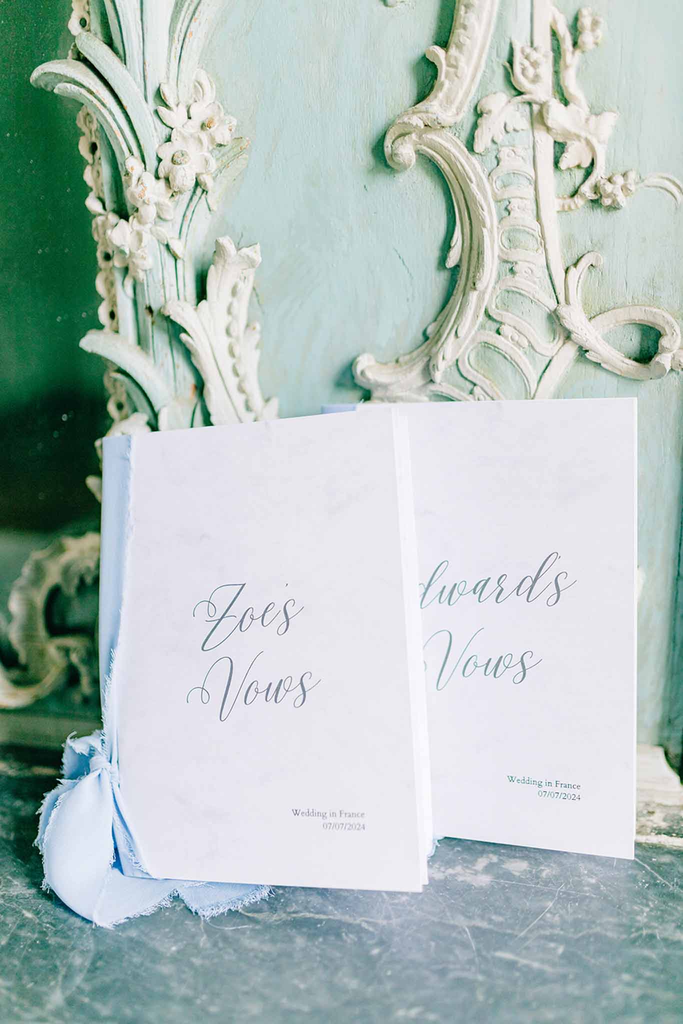 Two cream vow booklets in blue-gray script with pale blue ribbon against mint green baroque plaster molding
